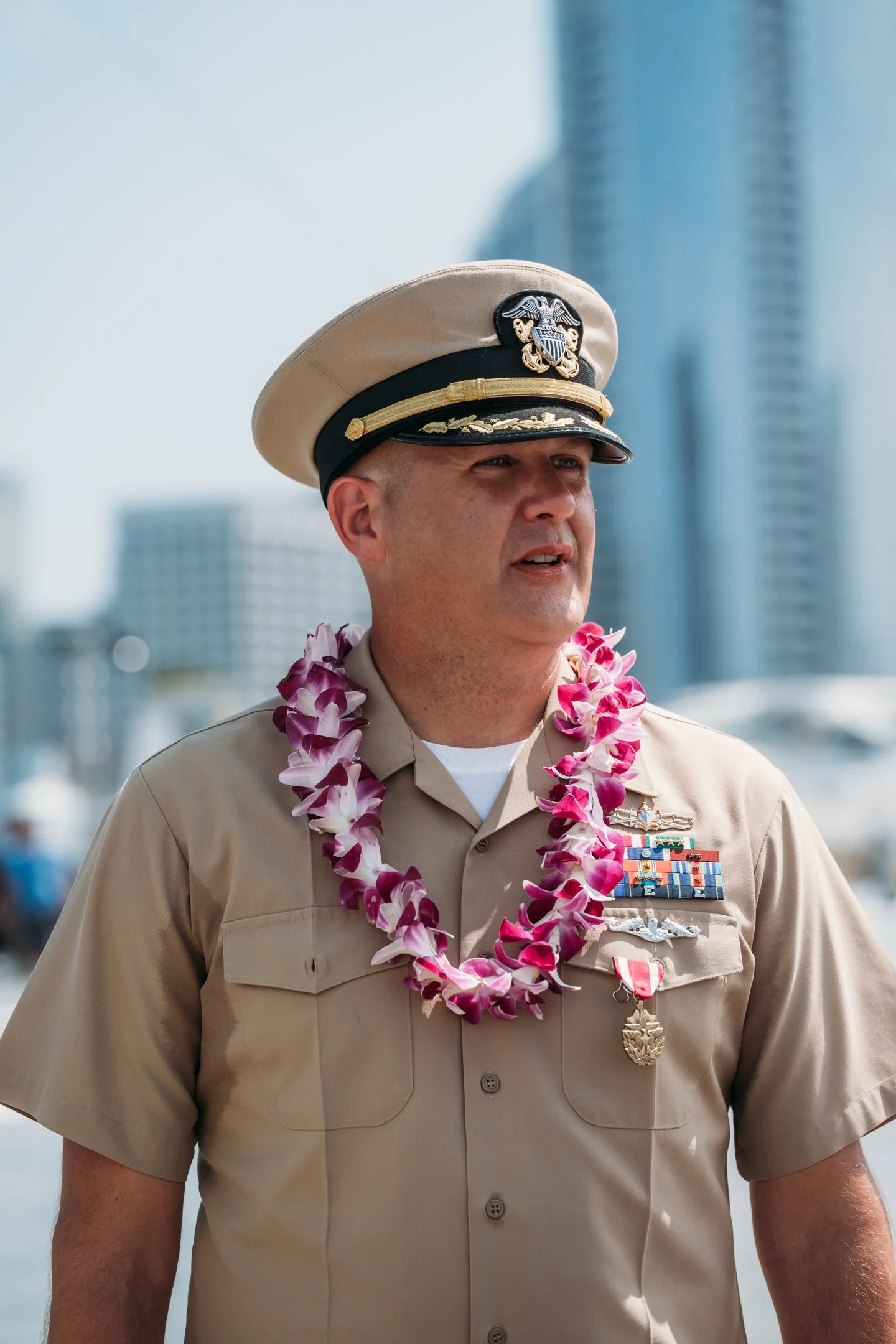A man in a U.S. Navy uniform with medals, a lei around his neck, and a hat with a Navy insignia, standing outdoors with city buildings in the background.