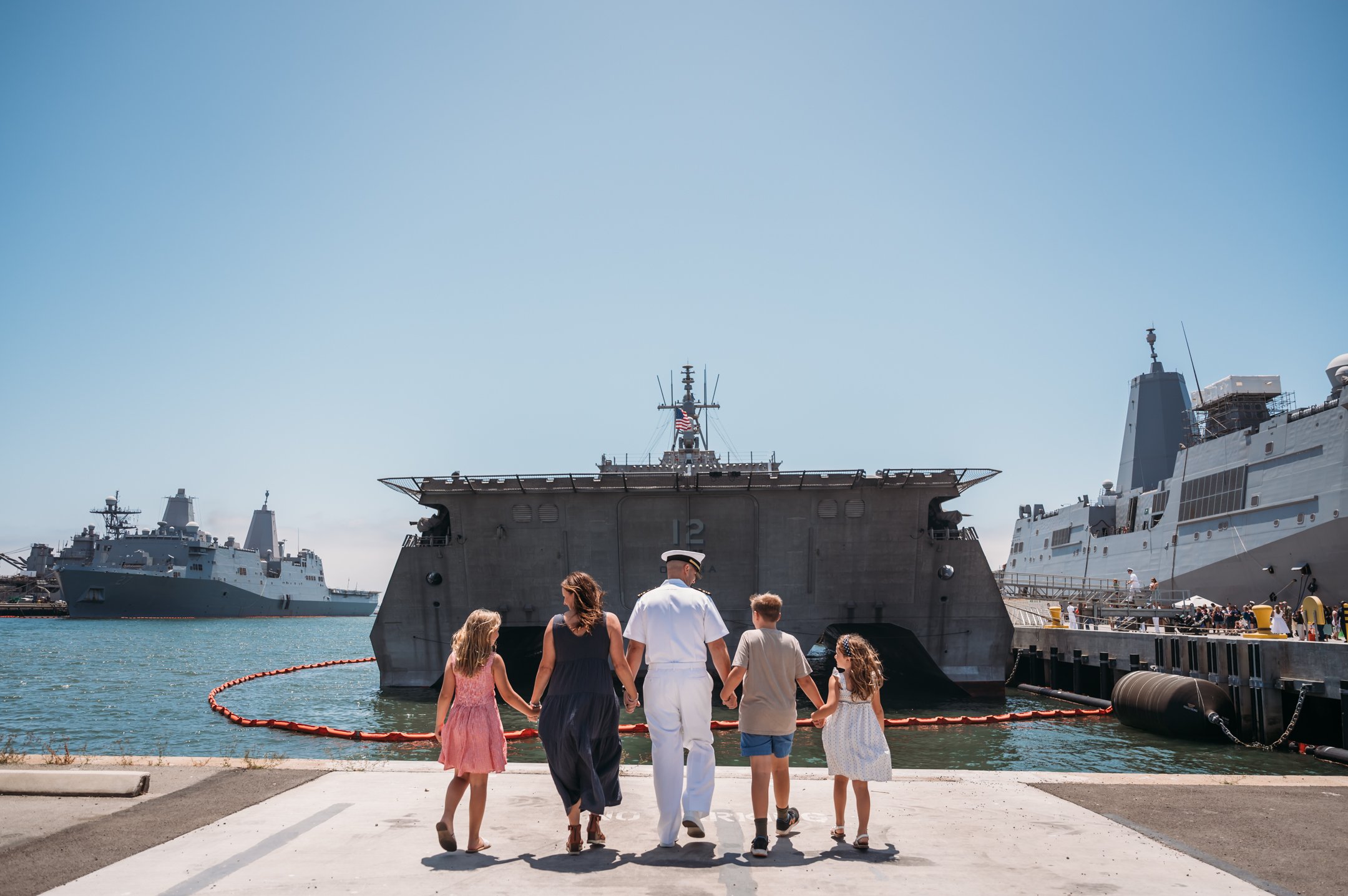 Family, including a woman, a man in a sailor uniform, and three children, walking hand in hand toward large military ships docked at a harbor on a sunny day. Alisha Mowry Photography Military Photographer San Diego