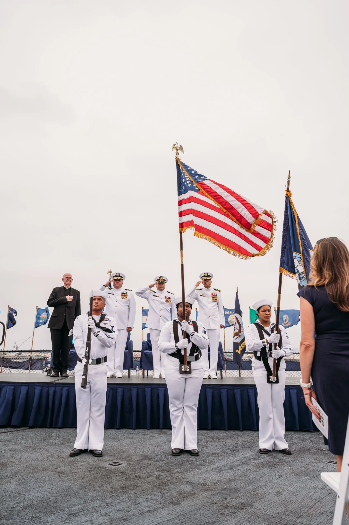 A military ceremony on a ship's deck with sailors in uniform holding flags, including the American flag, while a woman in civilian clothing watches. Alisha Mowry Photography Military Photographer San Diego
