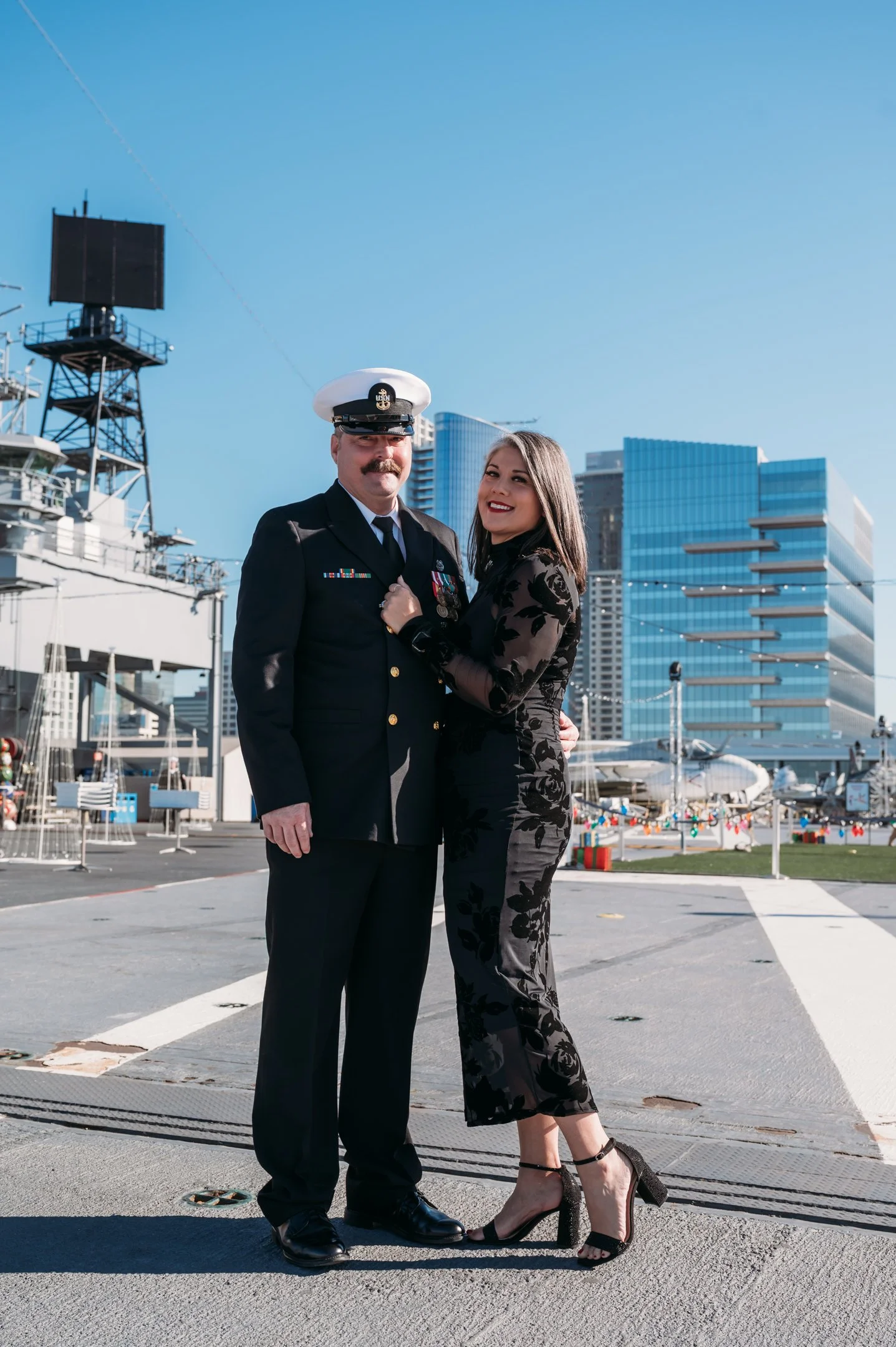 A man in a navy navy uniform with medals and a white hat stands beside a woman in a black floral dress and high heels, both smiling, outdoors in an urban setting with a modern glass building and boats in the background. uss midway military retirement