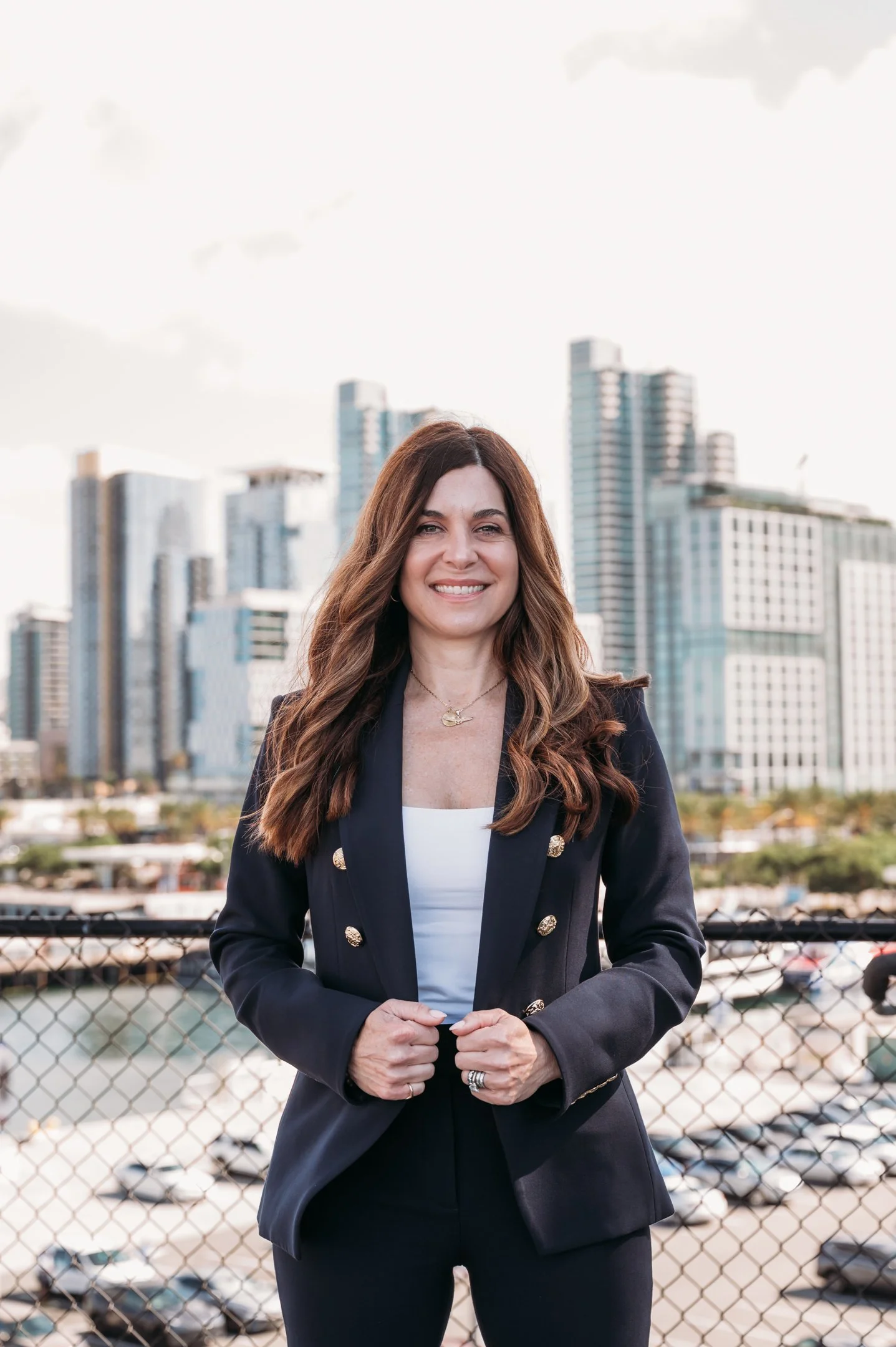 A woman with long, wavy brown hair, smiling, wearing a dark blazer with gold buttons over a white top, standing outdoors near a cityscape with tall buildings and parked cars in the background. Alisha Mowry Photography Military, Brand, and Portrait Ph