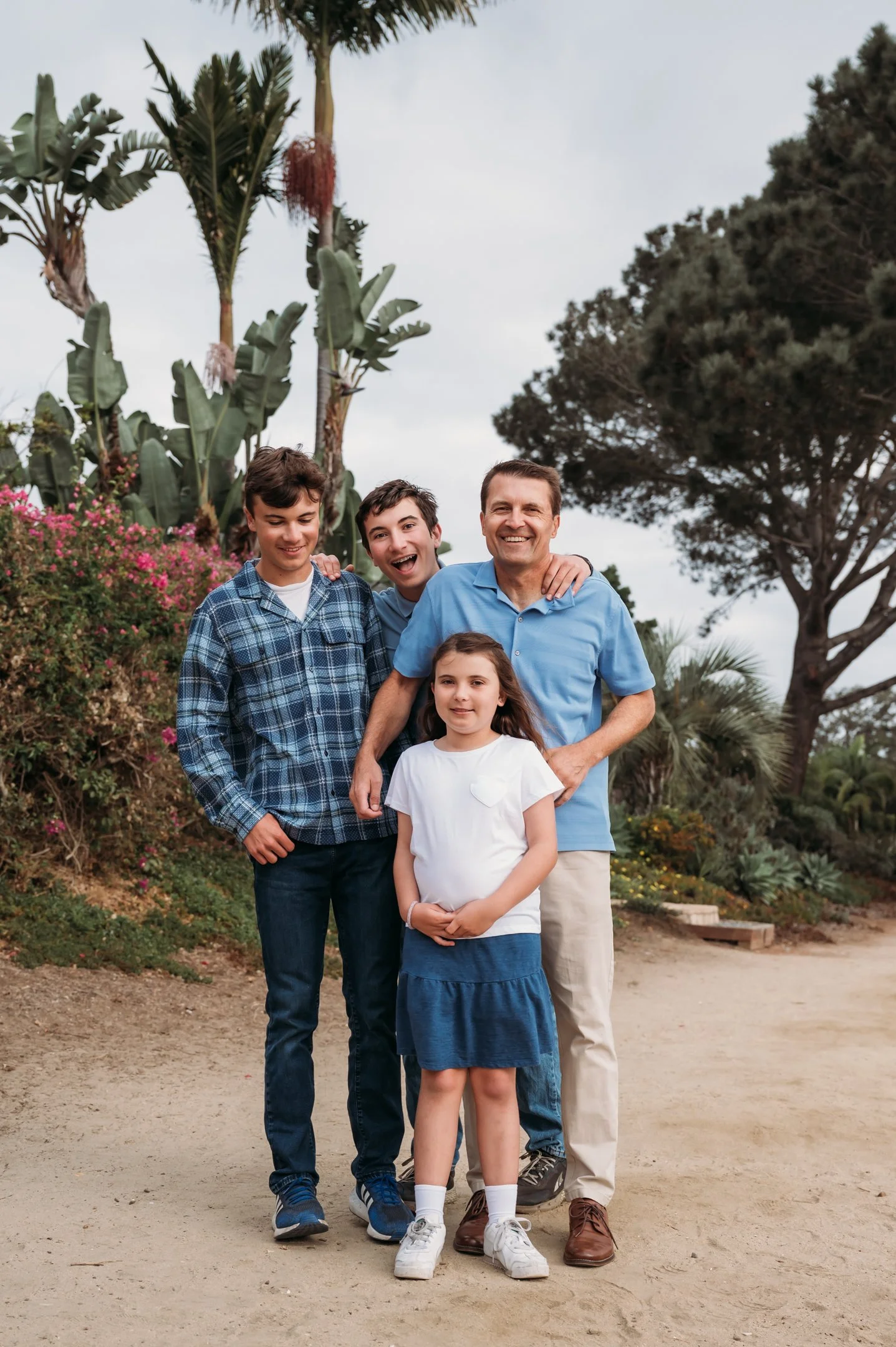 A smiling family of five standing outdoors on a dirt path with greenery and tall plants behind them. Alisha Mowry Photography Military, Brand, and Portrait Photographer San Diego CA