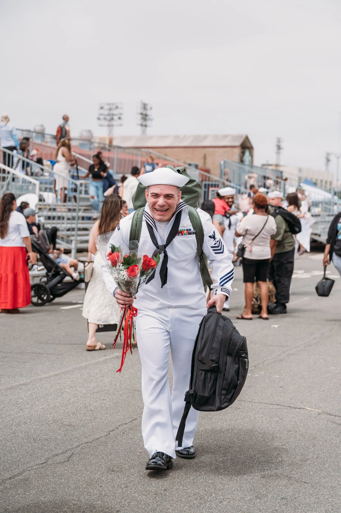 A smiling man in a U.S. Navy uniform holding a bouquet of roses walks across a schoolyard or stadium with people in the background. San Diego Military Homecoming Photograph