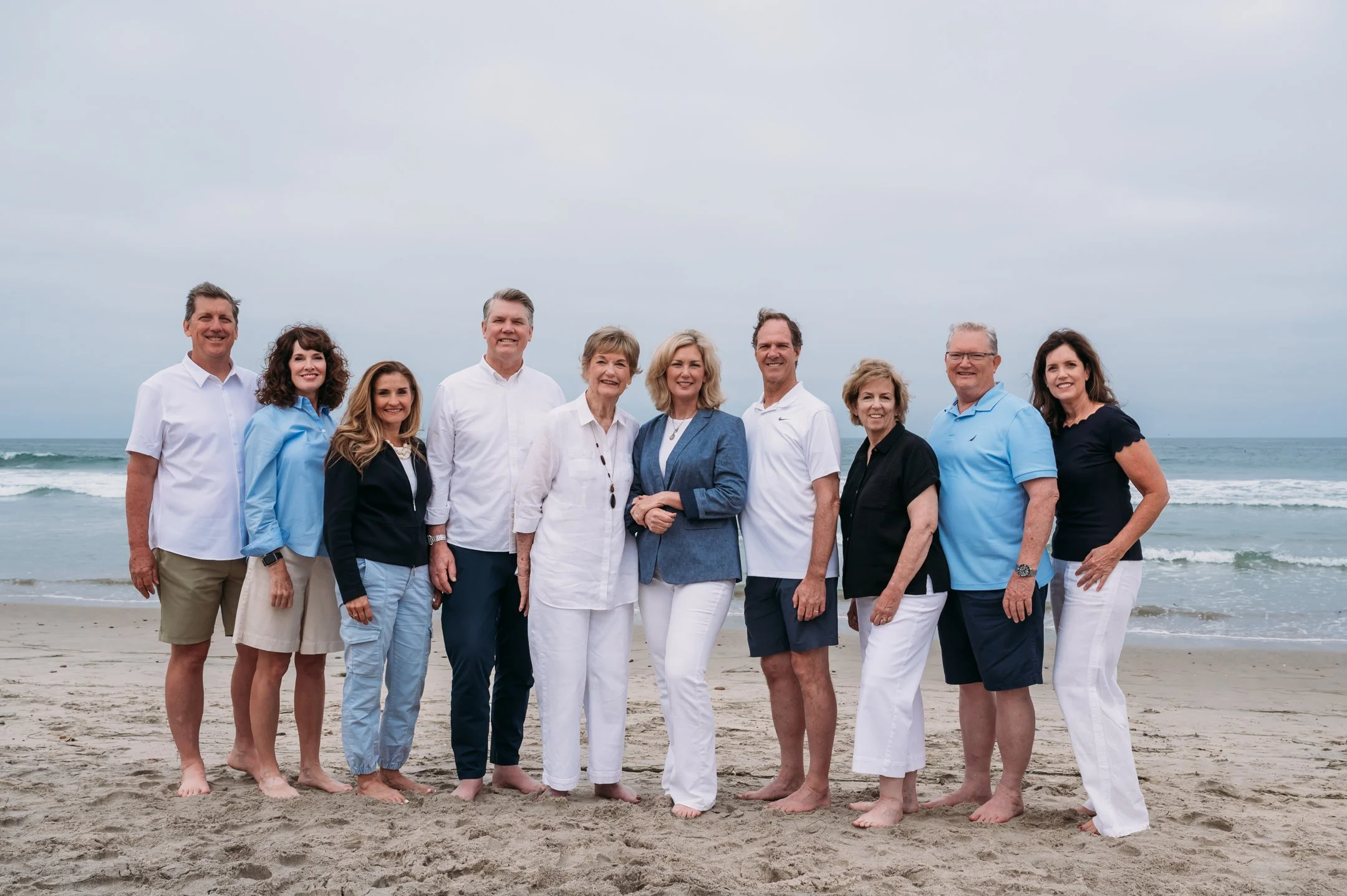 Group of ten adults on a beach, standing on sand with ocean waves and cloudy sky in the background.