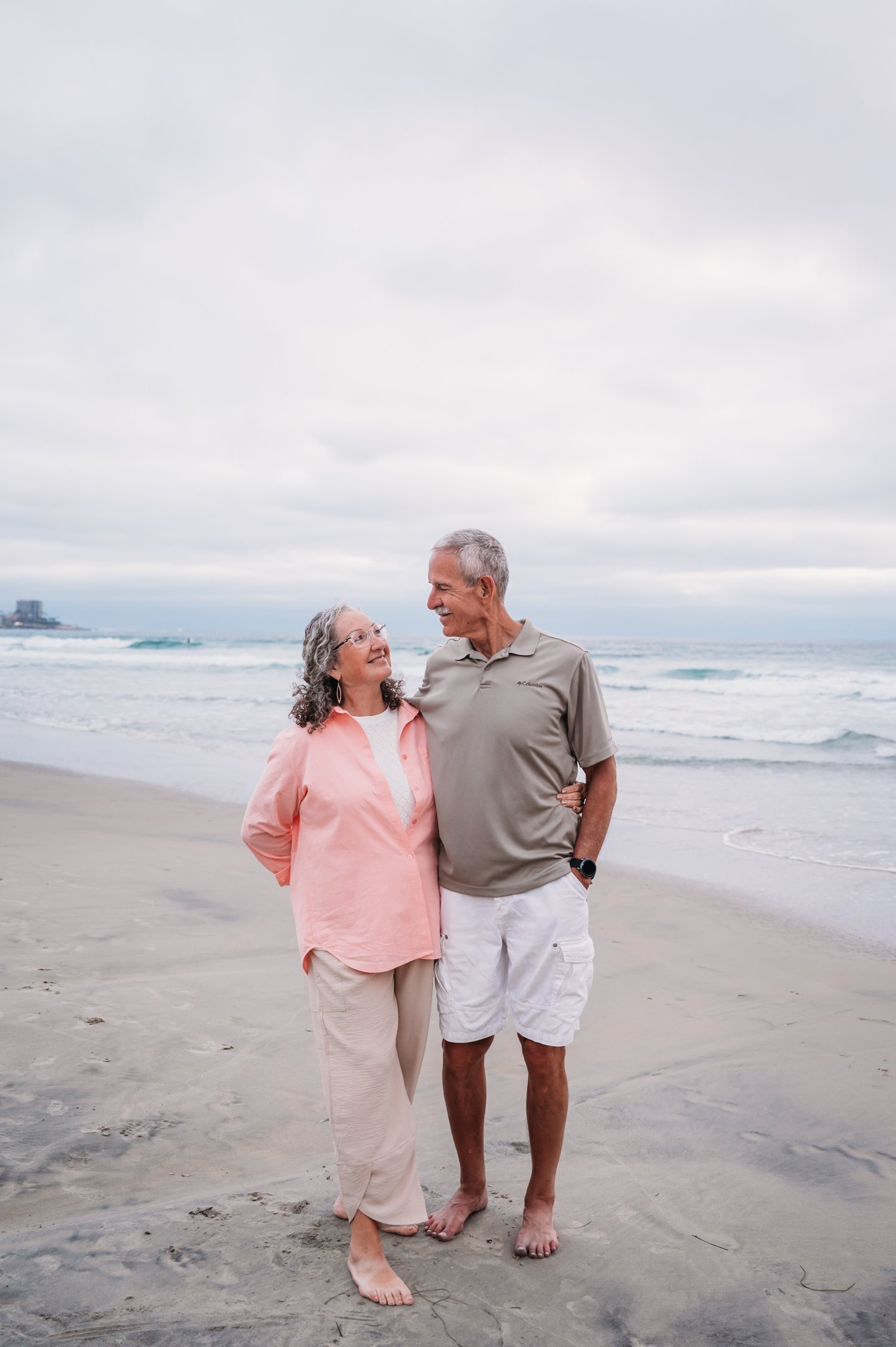 Older couple smiling and walking barefoot on a beach with cloudy sky and ocean waves in the background.