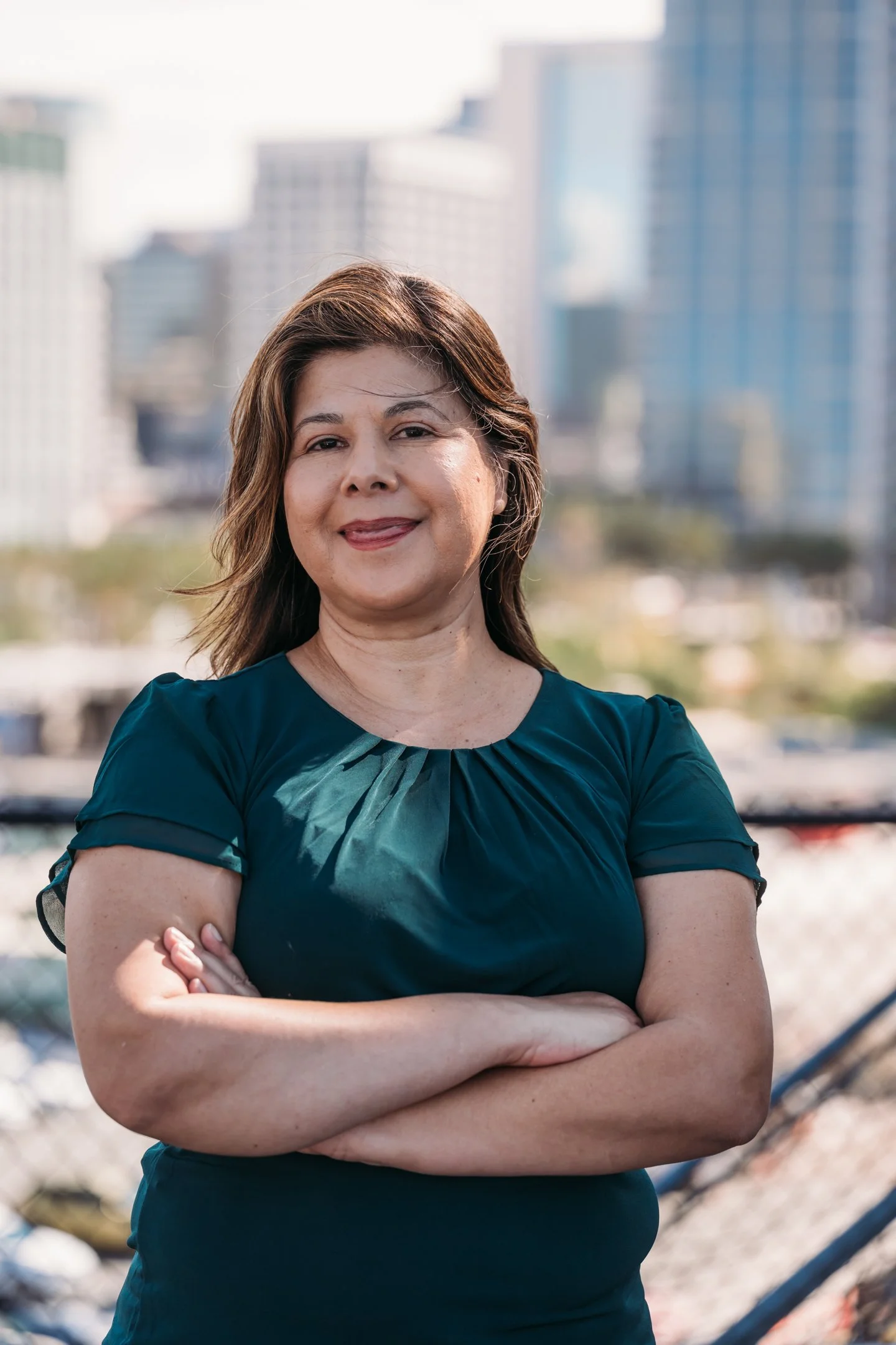 A woman with shoulder-length brown hair standing outdoors with arms crossed, smiling, with a city skyline in the background. Alisha Mowry Photography Military, Brand, and Portrait Photographer San Diego CA