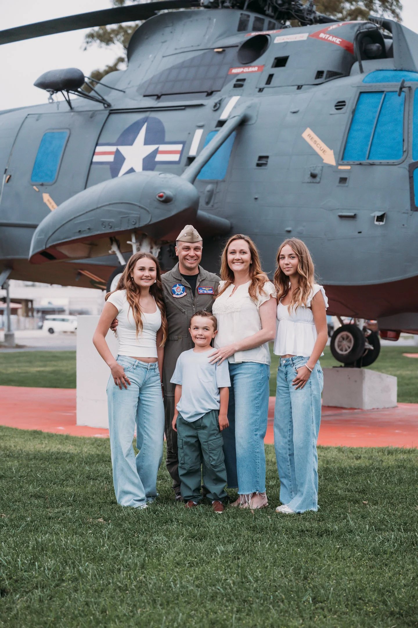 Family of five, including a man in a military uniform, posing in front of a military helicopter outdoors on a grassy area.