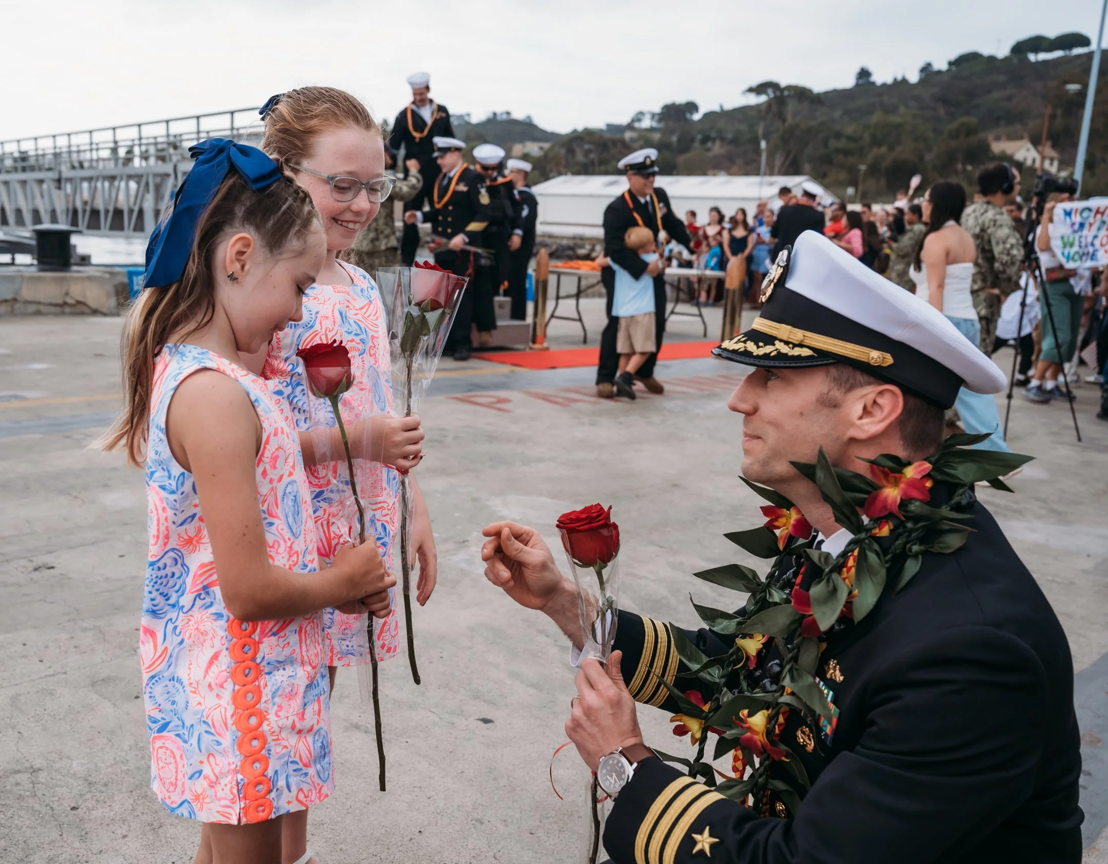 A man in a military uniform kneels to present flowers to two young girls holding roses in plastic wrap during a graduation or celebration ceremony. The girls wear colorful dresses with blue ribbons in their hair, and the man wears a white cap decorat