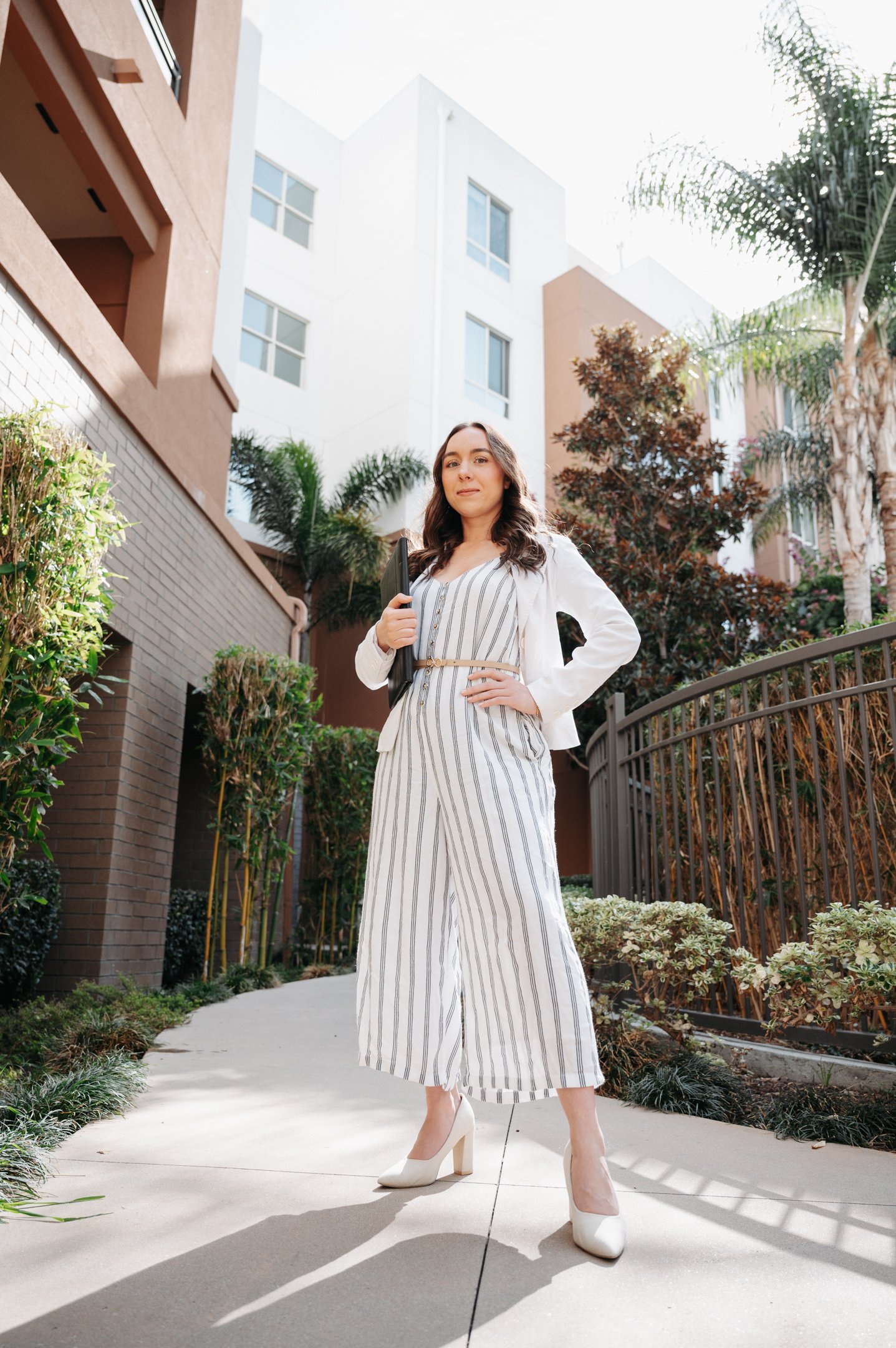 A woman in a striped jumpsuit and white blazer standing outdoors on a sidewalk, holding a folder, with apartment buildings and trees in the background. Alisha Mowry Photography Military, Brand, and Portrait Photographer San Diego CA