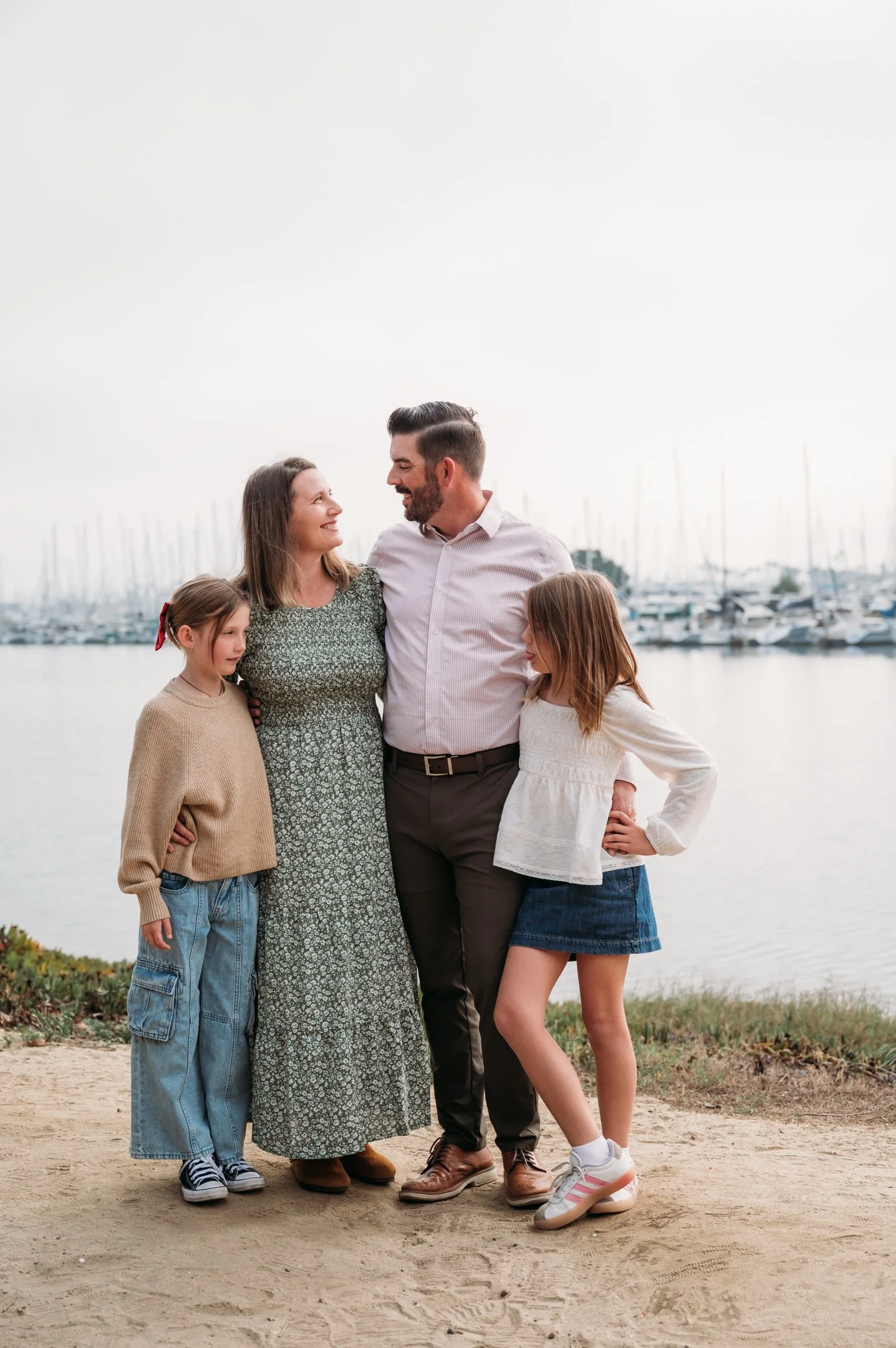 A family of five standing on a dirt path near a body of water with boats in the background, smiling and looking at each other. Alisha Mowry Photography Military, Brand, and Portrait Photographer San Diego CA
