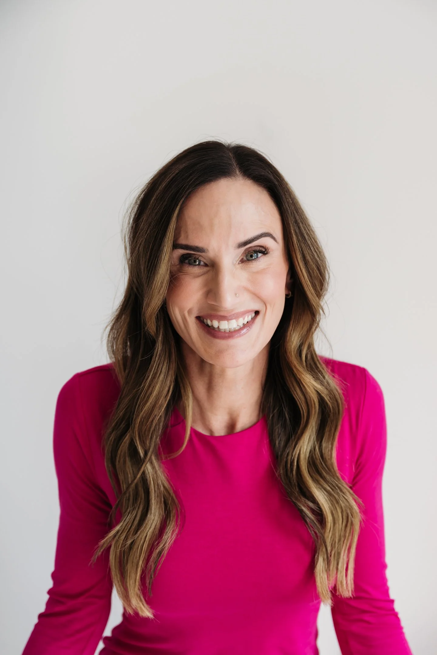 A woman with long brown hair, wearing a bright pink top, smiling at the camera against a plain white background. Alisha Mowry Photography Military, Brand, and Portrait Photographer San Diego CA