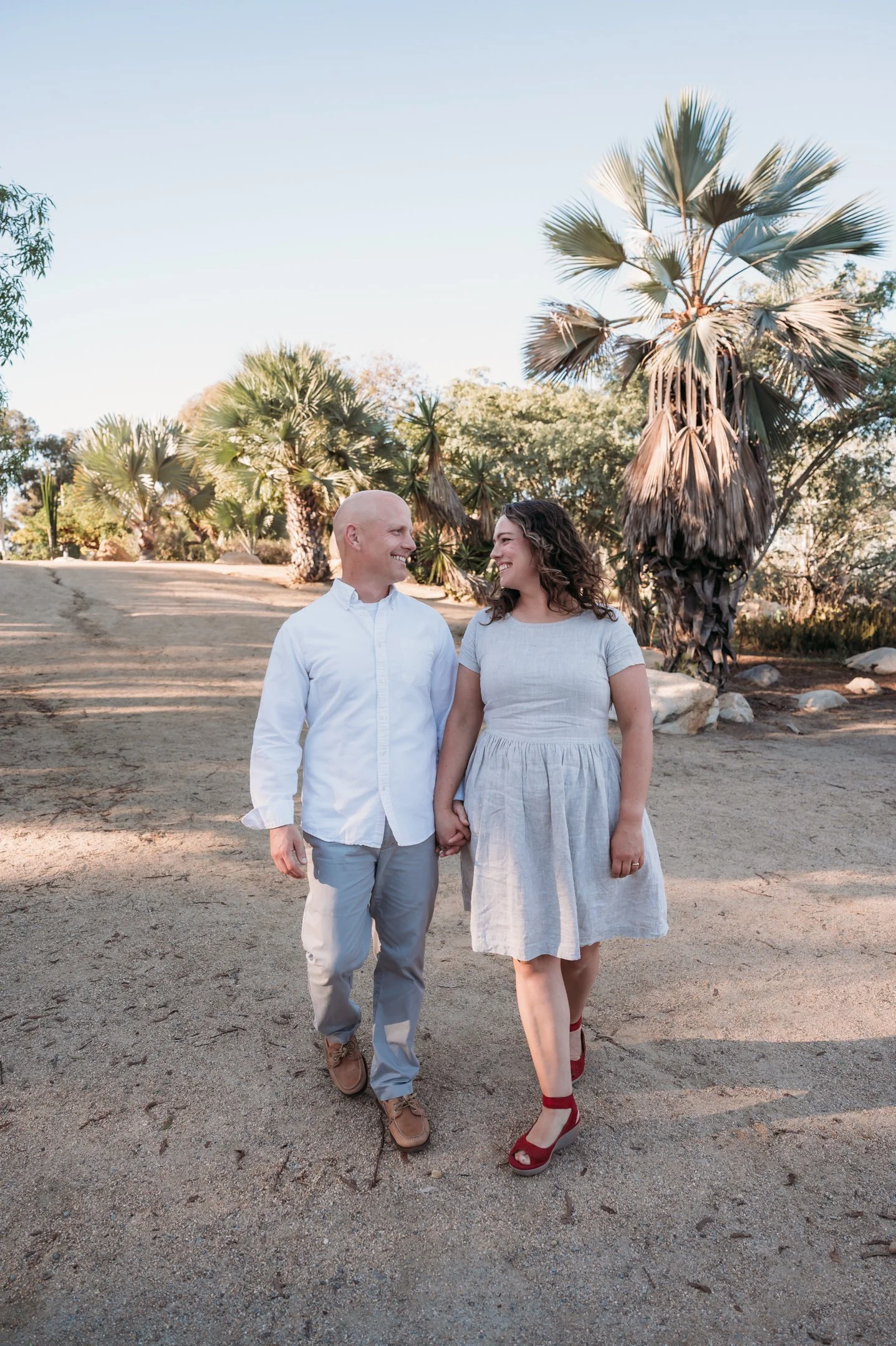 A couple holding hands and smiling at each other while walking outdoors in a sunny, desert-like area with palm trees and rocks in the background. Alisha Mowry Photography Military, Brand, and Portrait Photographer San Diego CA