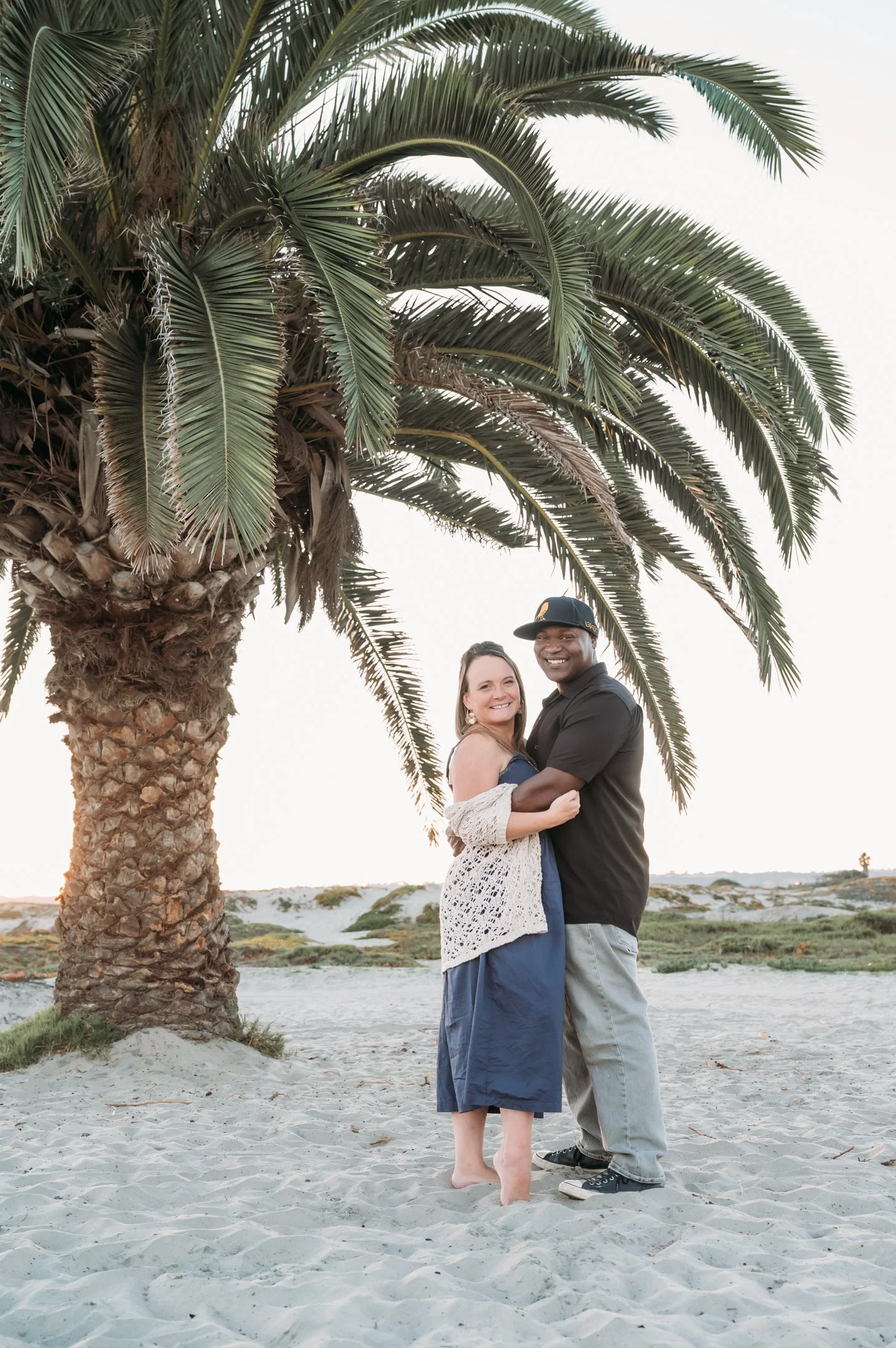 A smiling couple standing on a sandy beach next to a large palm tree, with the setting sun in the background. Alisha Mowry Photography Military, Brand, and Portrait Photographer San Diego CA
