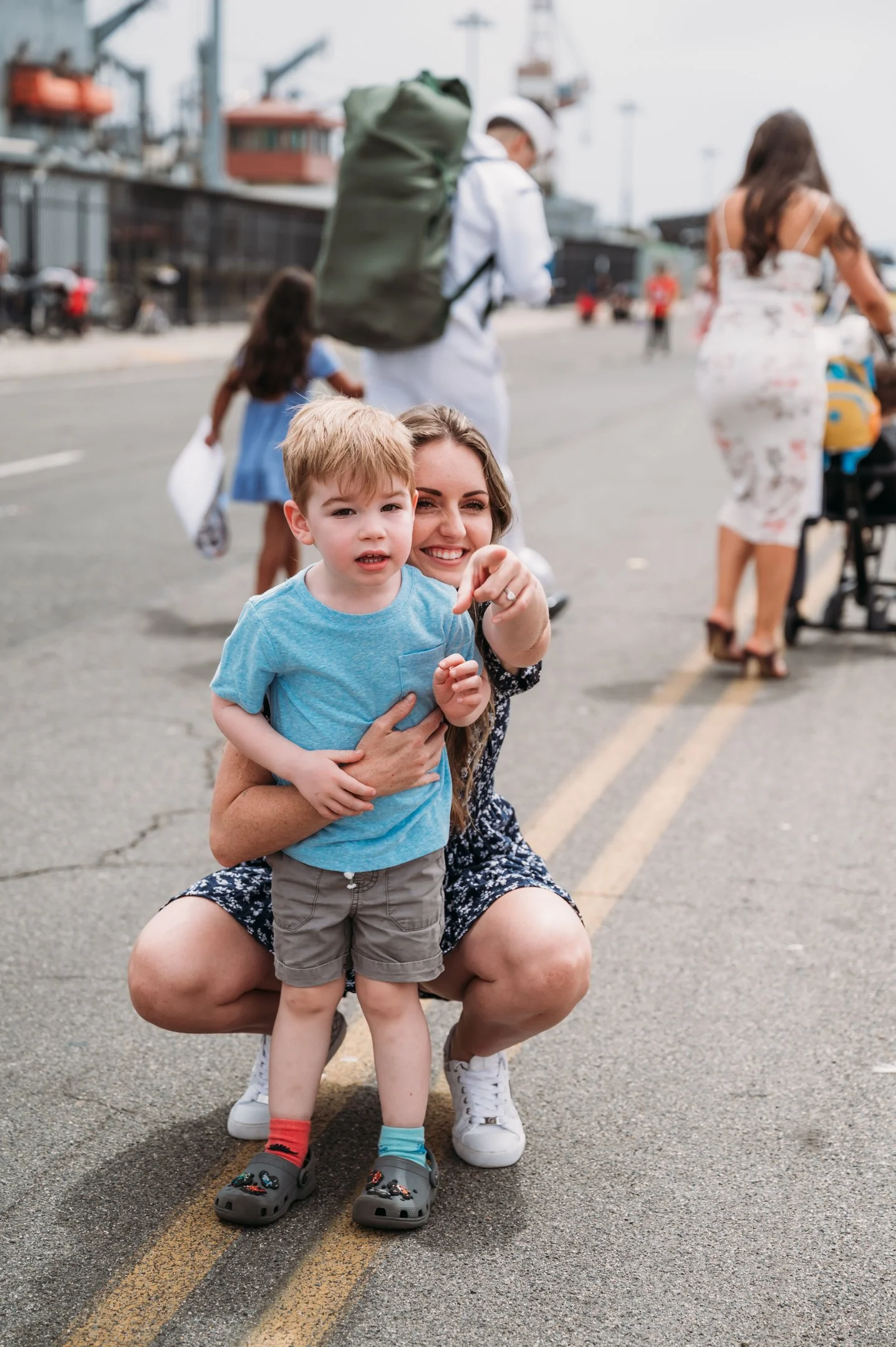 A woman crouching in the street, holding a young boy in front of her, both looking at the camera. The street is busy with people and docked ships in the background. San Diego Military Homecoming Photograph