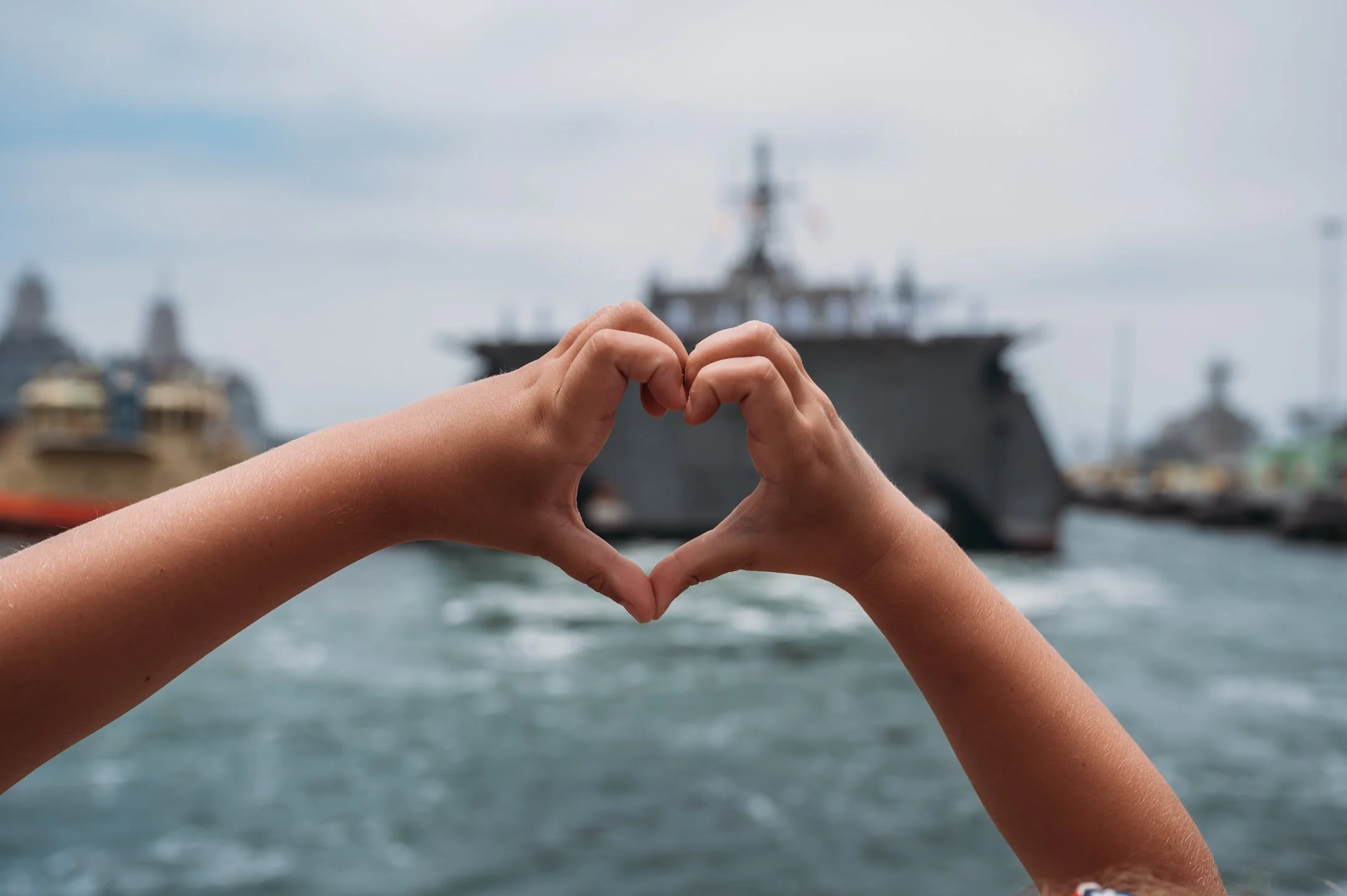Hands forming a heart shape in front of a large navy ship in a harbor. Alisha Mowry Photography Military Photographer San Diego