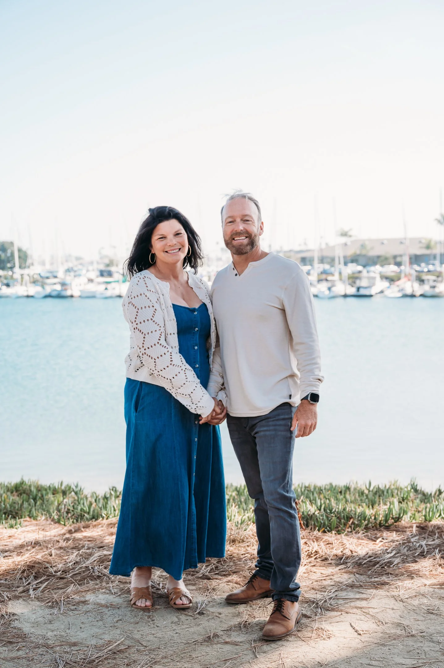 A smiling couple holding hands in front of a marina with sailboats, standing on a dirt path with grass, under a clear sky. Alisha Mowry Photography Military, Brand, and Portrait Photographer San Diego CA