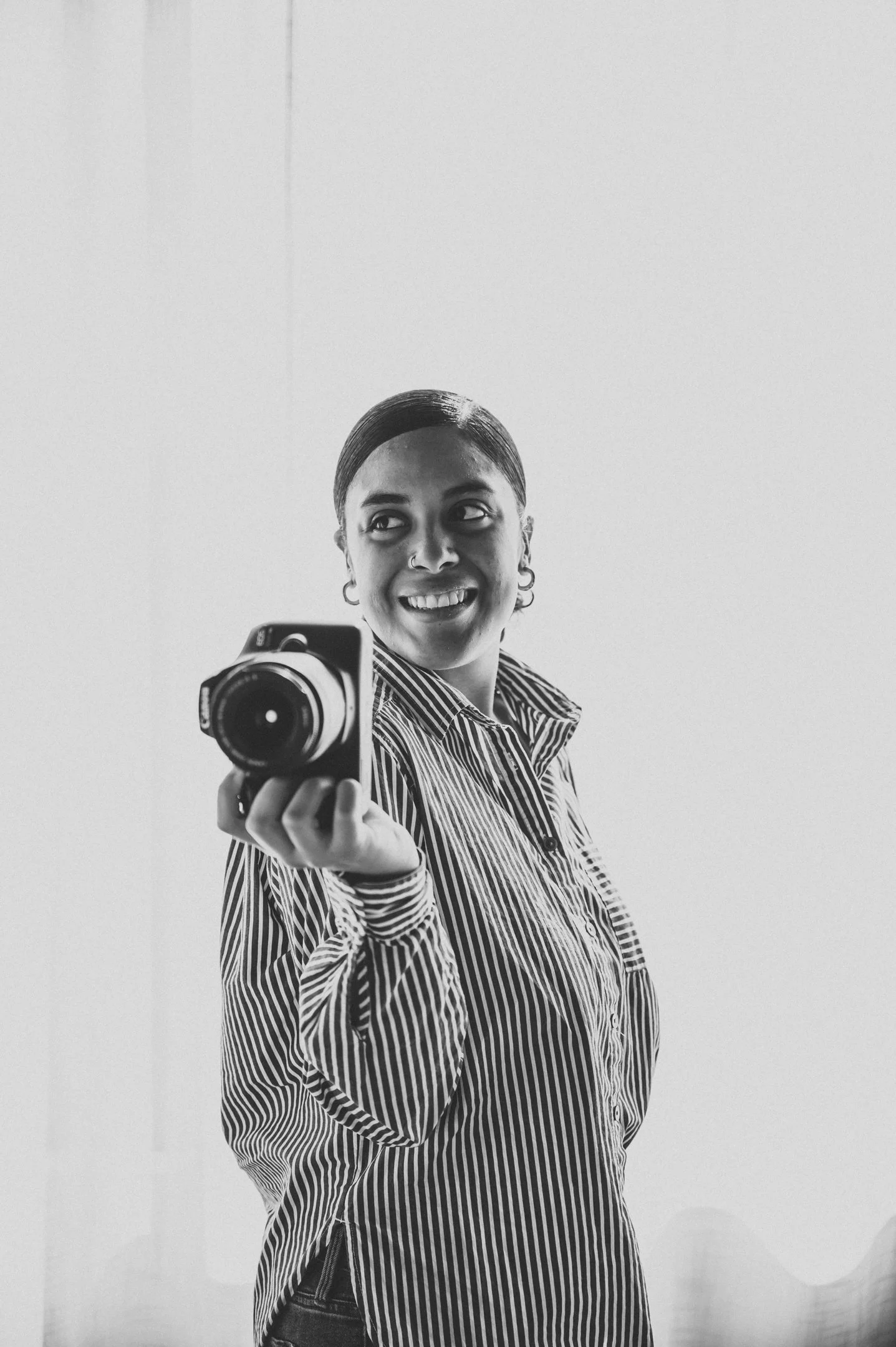 Black-and-white photo of a woman taking a selfie in a mirror, holding a camera with her right hand, smiling while wearing a striped shirt, with her hair slicked back. Alisha Mowry Photography Military, Brand, and Portrait Photographer San Diego CA