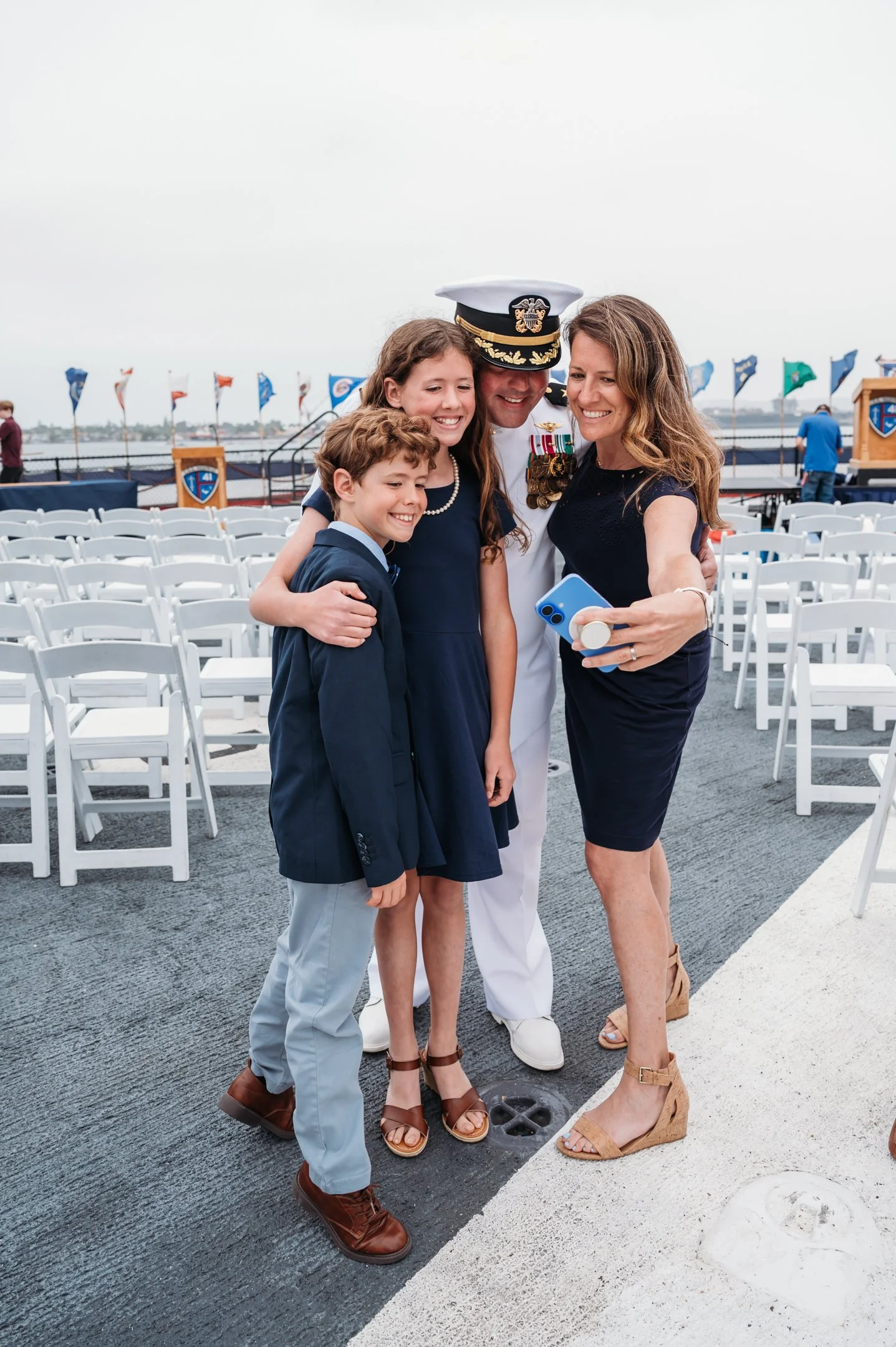 A woman taking a selfie with a man in a Navy uniform and two children on a boat deck, with white chairs and flags in the background. Alisha Mowry Photography Military Photographer San Diego