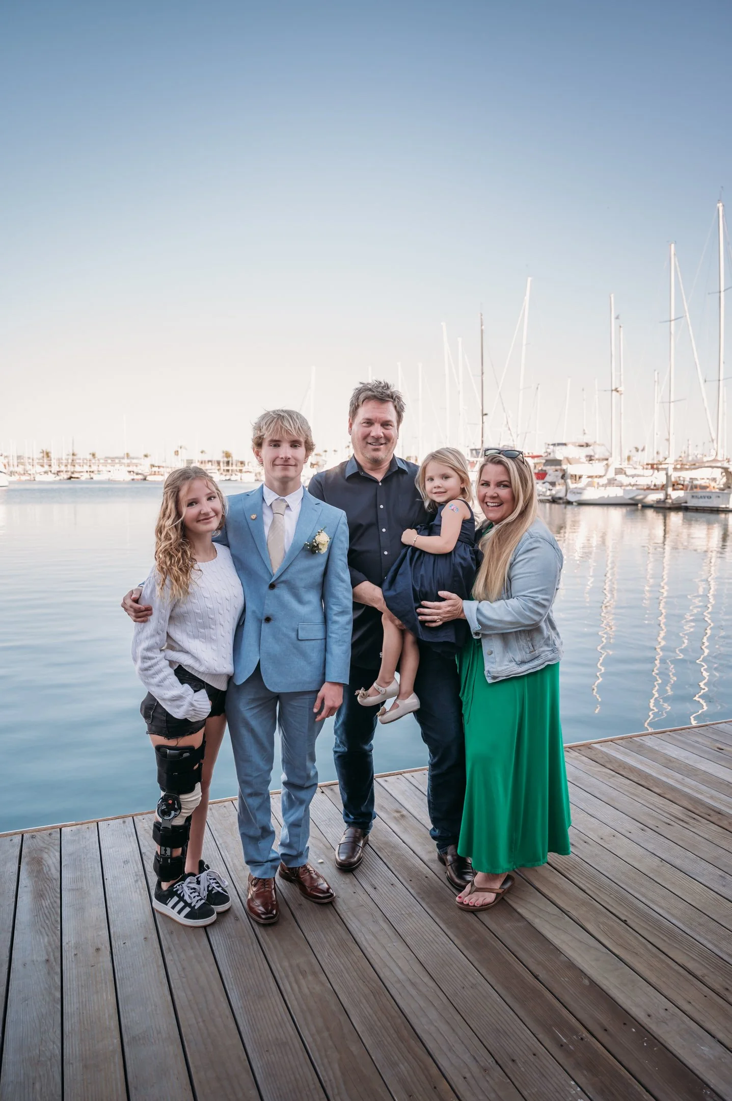 A family of five standing on a wooden dock by the water with sailboats in the background. They are smiling and dressed casually. Alisha Mowry Photography Military Photographer Sa