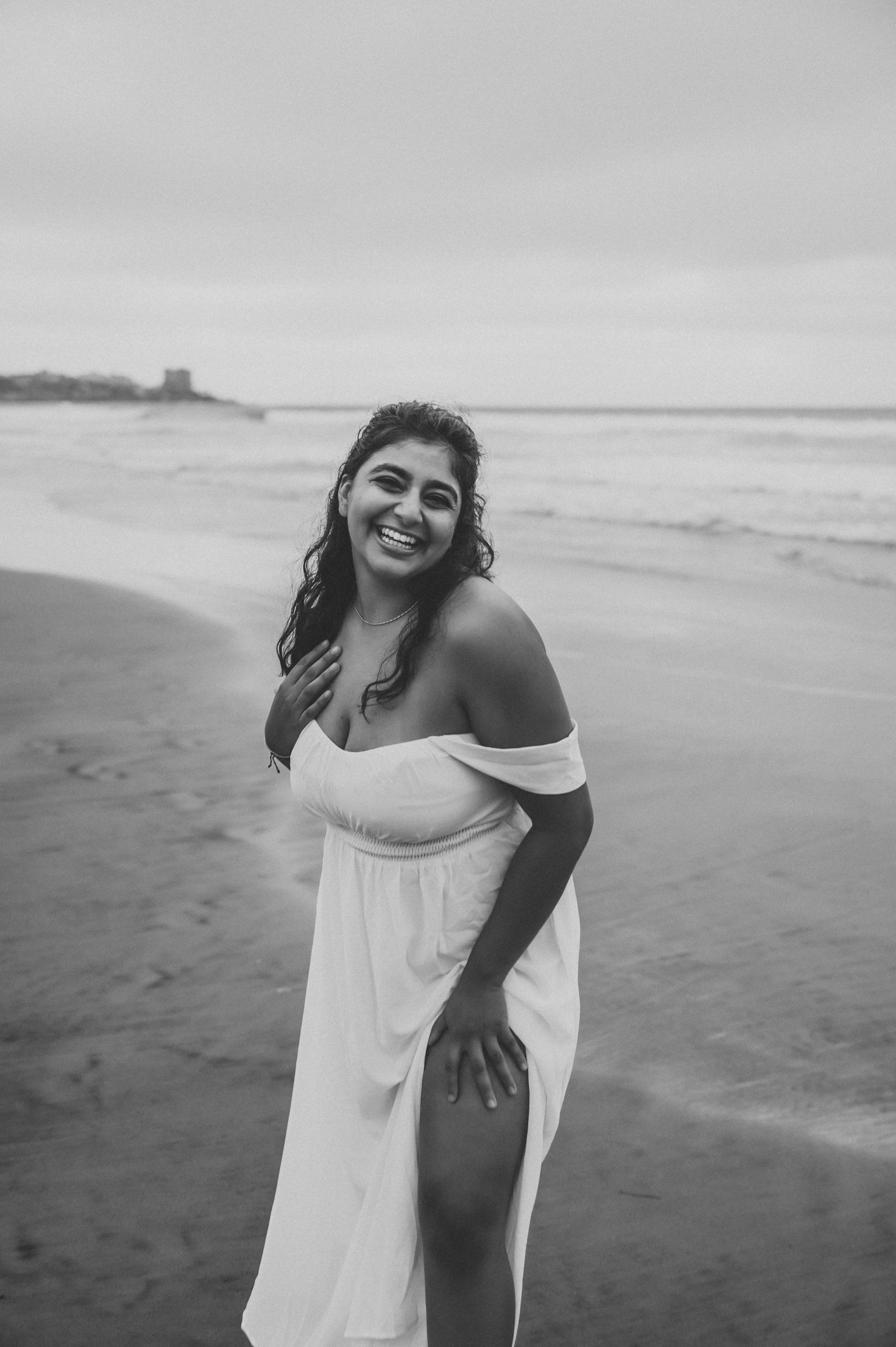 A smiling woman with long, curly hair stands on the beach in a white off-shoulder dress, with the ocean and shoreline in the background. Alisha Mowry Photography Military, Brand, and Portrait Photographer San Diego CA