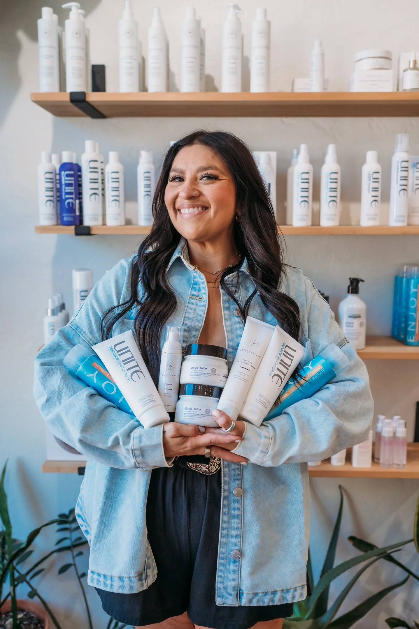 A woman smiling and holding various skincare and beauty products, standing in front of shelves filled with similar products in a retail setting. Alisha Mowry Photography Military, Brand, and Portrait Photographer San Diego CA