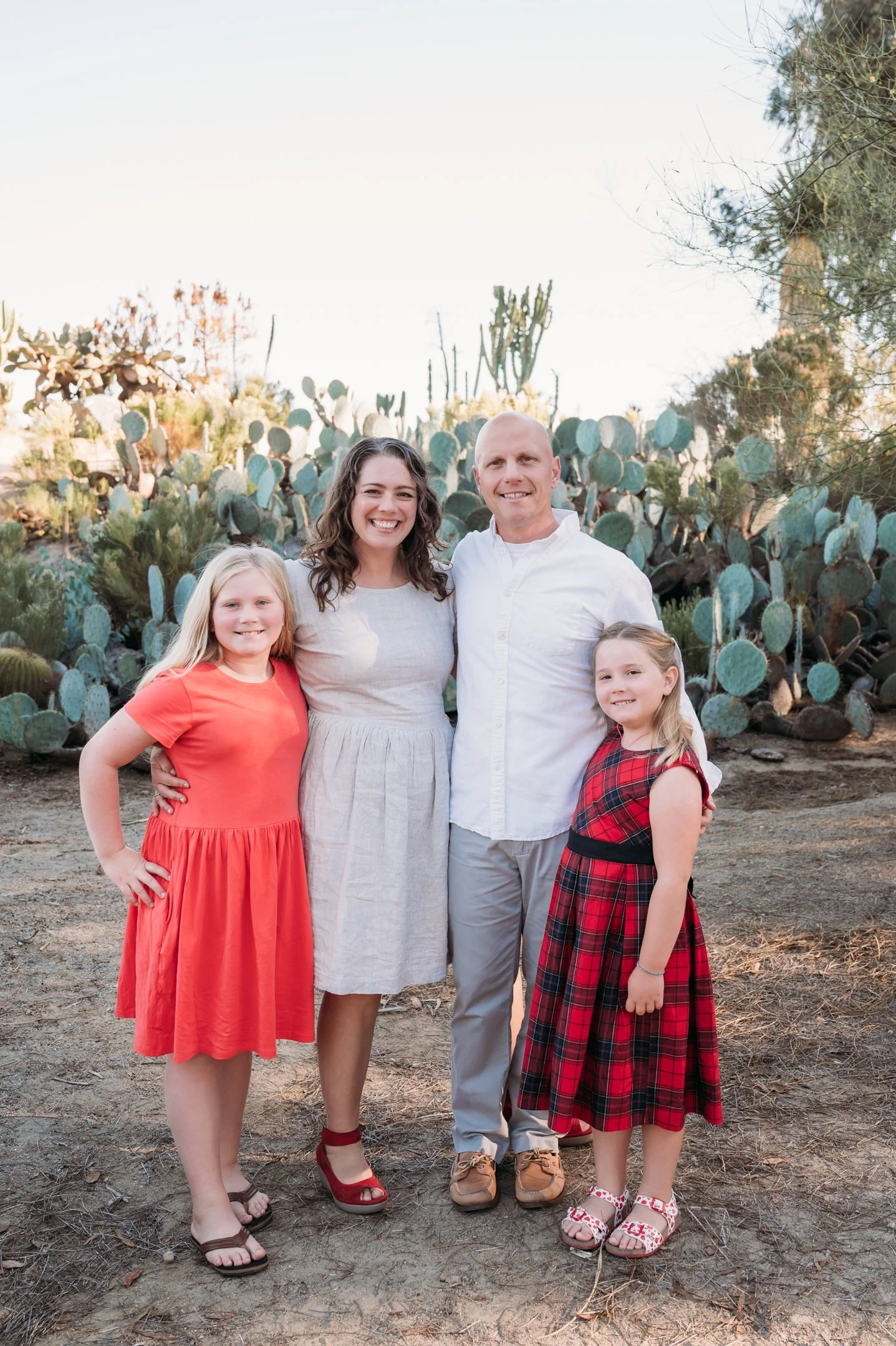 Family of four standing outdoors in front of cactus plants, smiling, with two young girls and a man and woman, dressed casually, on a dirt ground during daytime. Alisha Mowry Photography Military, Brand, and Portrait Photographer San Diego CA