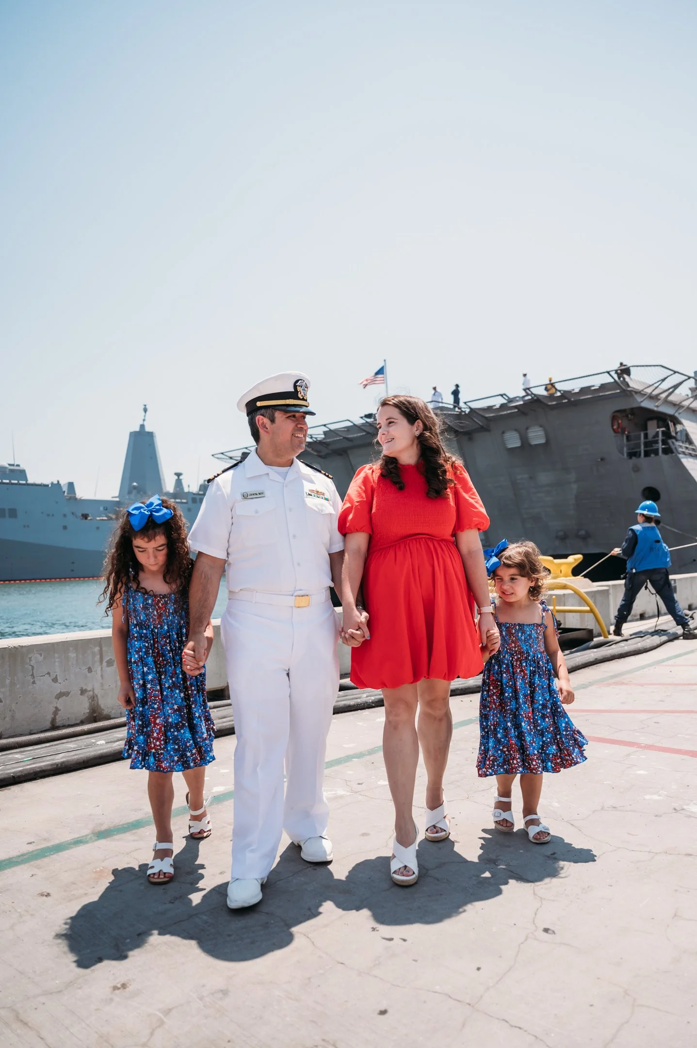 A family walking hand-in-hand along a dock on a sunny day, with a naval ship in the background. The father is dressed in a white naval uniform, the mother in a red dress, and the two daughters in blue floral dresses with large blue bows.