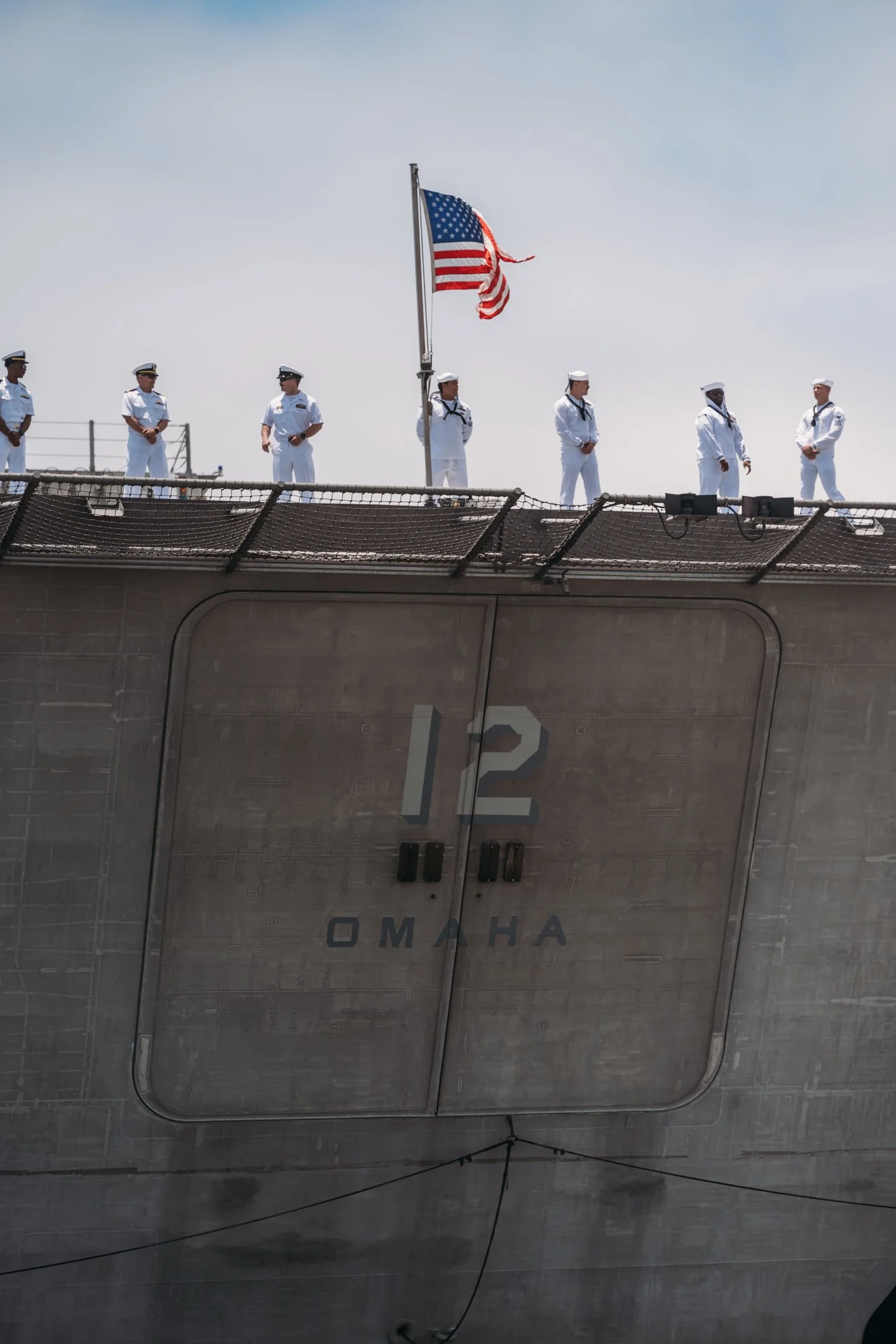 Military personnel in white uniforms stand on a ship's deck near an American flag, with a large number 12 and the word 'Omaha' visible on the side of the vessel. San Diego Military Homecoming Photograph