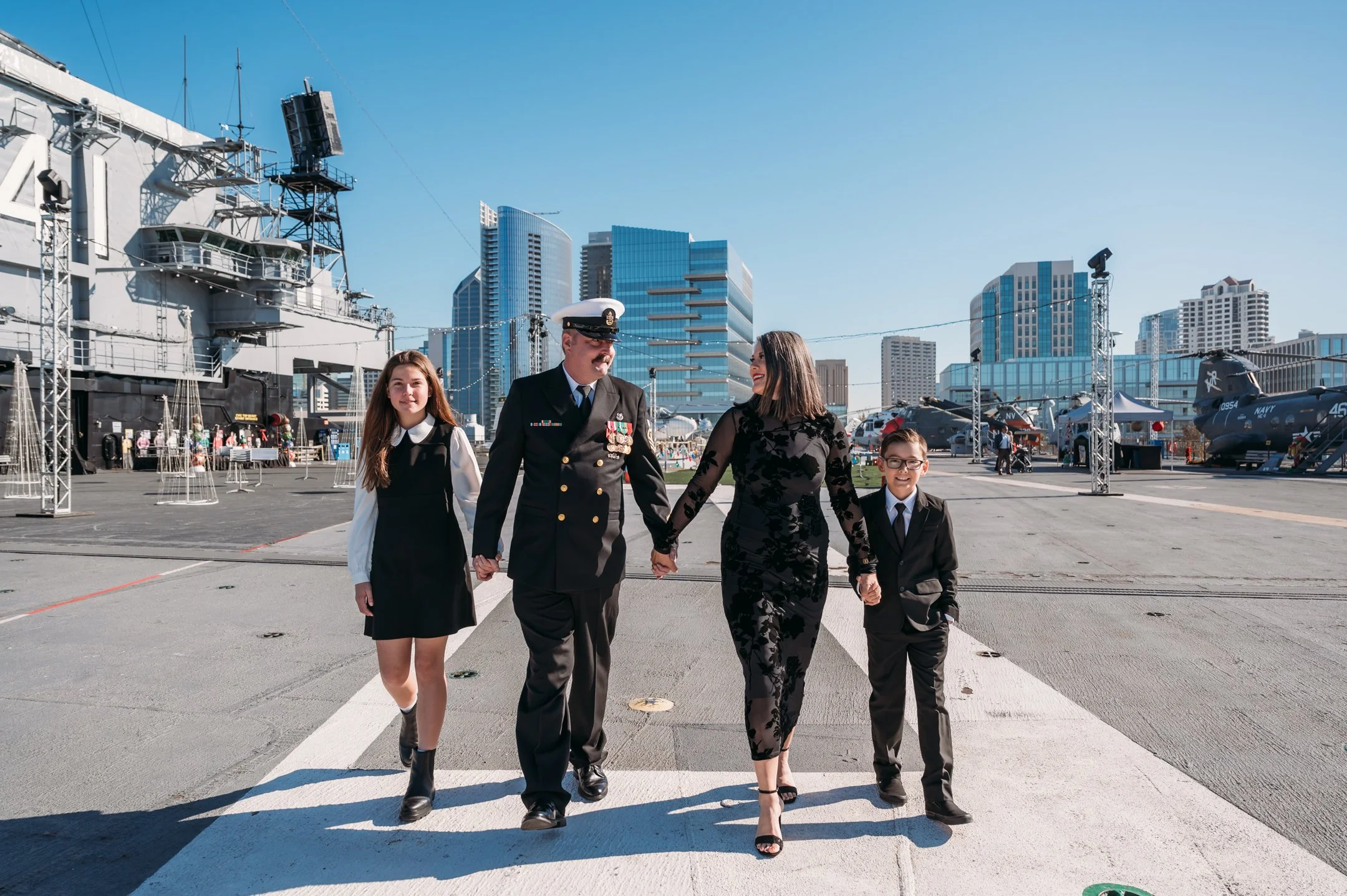 Family walking hand-in-hand on an aircraft carrier at a naval museum, with aircraft and san diego skyline in background. uss midway military retirement ceremony 