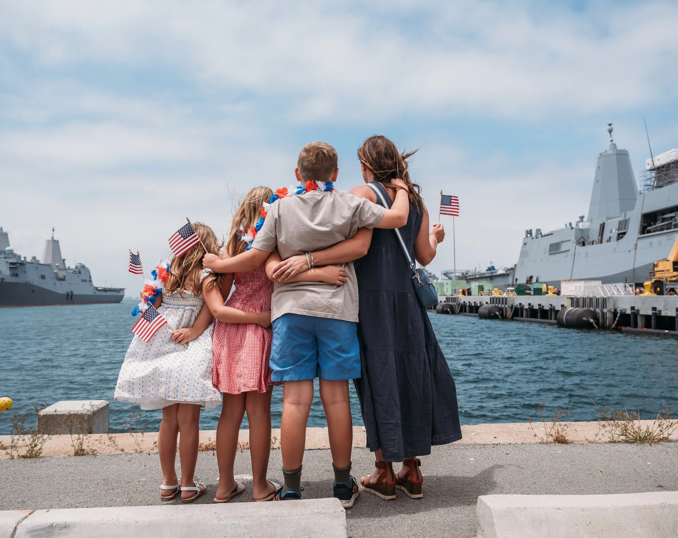 A family of four, children and a woman, celebrating by a harbor with navy ships in the background, holding American flags and wearing patriotic accessories. Alisha Mowry Photography Military Photographer San Diego