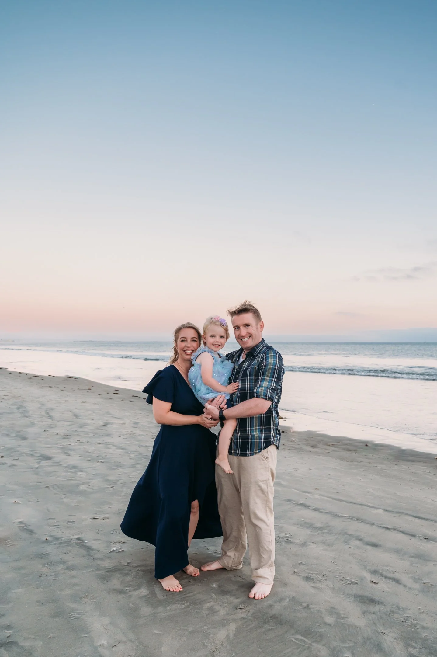 A family of three standing barefoot on the sandy beach at sunset, smiling and looking at the camera, with the ocean and pastel sky in the background. Alisha Mowry Photography Military, Brand, and Portrait Photographer San Diego CA