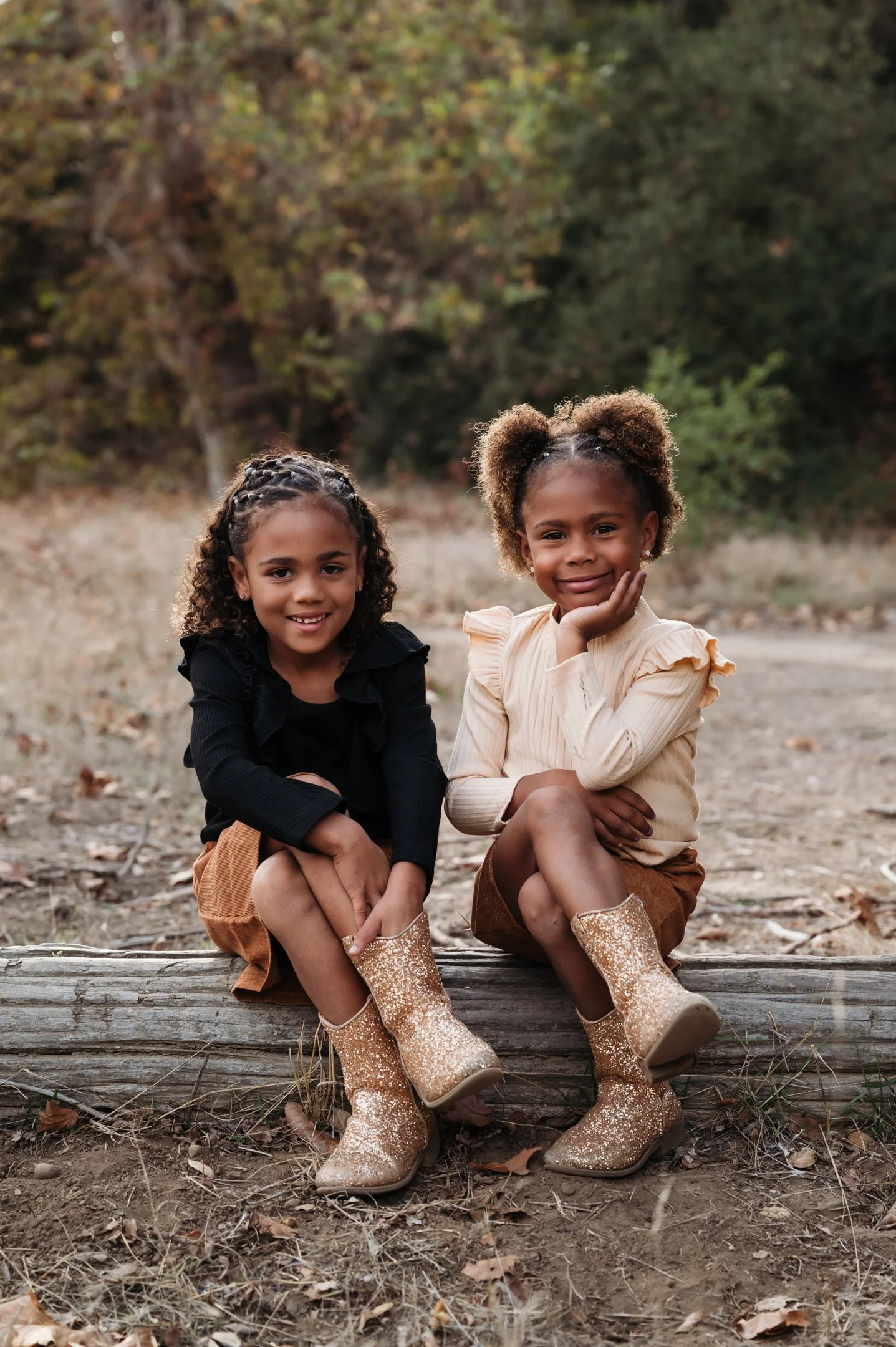 Two young girls sitting on a log outdoors with trees in the background, wearing boots and smiling. Alisha Mowry Photography Military, Brand, and Portrait Photographer San Diego CA