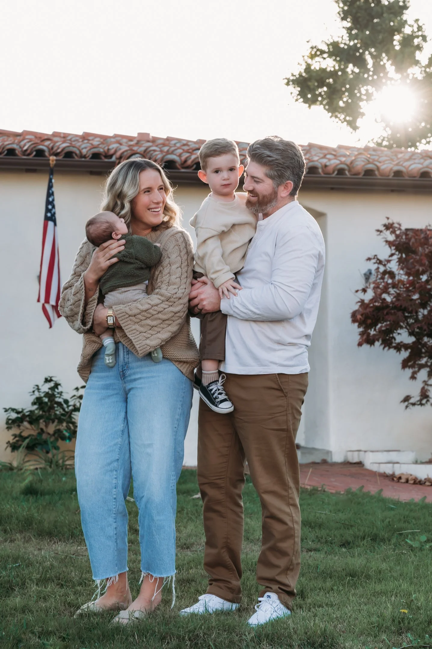 A family of four standing outdoors on a lawn, with a house and American flag in the background, sun shining behind a tree, smiling and holding children. Alisha Mowry Photography Military, Brand, and Portrait Photographer San Diego CA