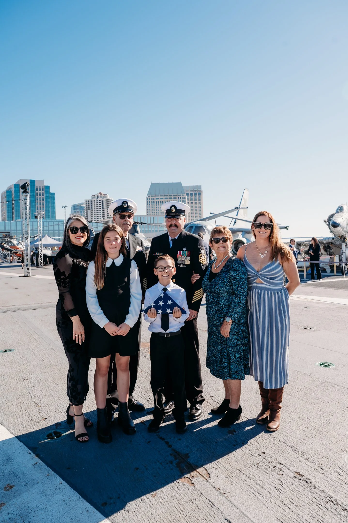 Group of people, including military personnel and civilians, standing on an outdoor aircraft carrier deck with fighter jets in the background on a sunny day. uss midway military promotion ceremony 