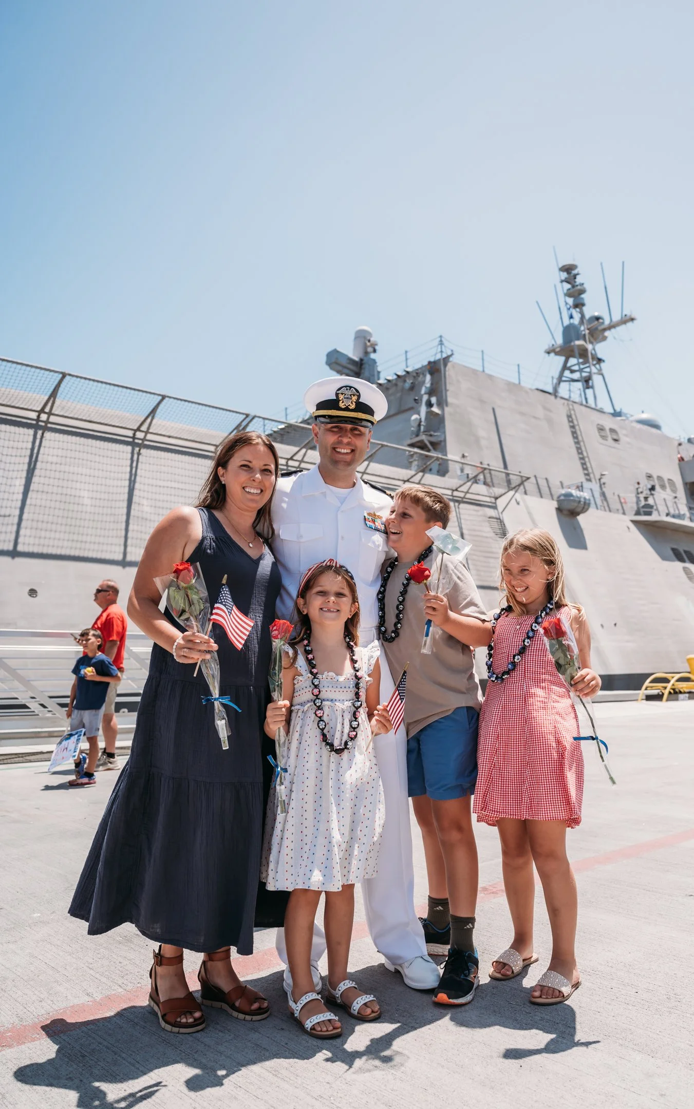 Family celebrating on a military ship's deck, with children holding US flags and roses, a naval officer in uniform, and people in the background. Alisha Mowry Photography Military Photographer San Diego