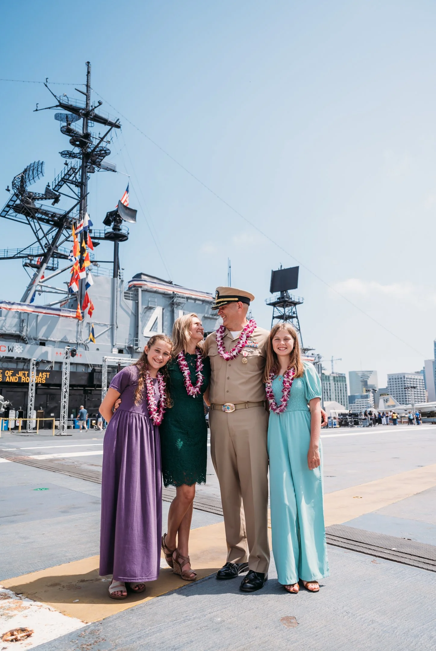 A family with two girls and an older woman wear leis and smile with a U.S. Navy officer in uniform at an aircraft carrier docked at a port, with city buildings in the background. uss midway military retirement ceremony 