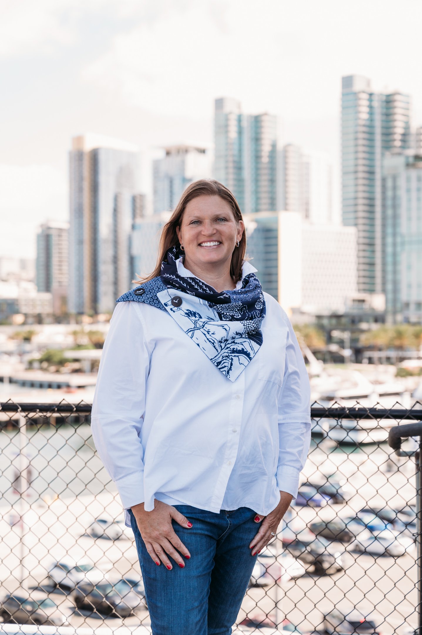 A woman standing outdoors at a rooftop or balcony, with a city skyline and marina in the background. She is smiling, wearing a white shirt, blue jeans, and a blue-and-white scarf. Alisha Mowry Photography Military, Brand, and Portrait Photographer Sa