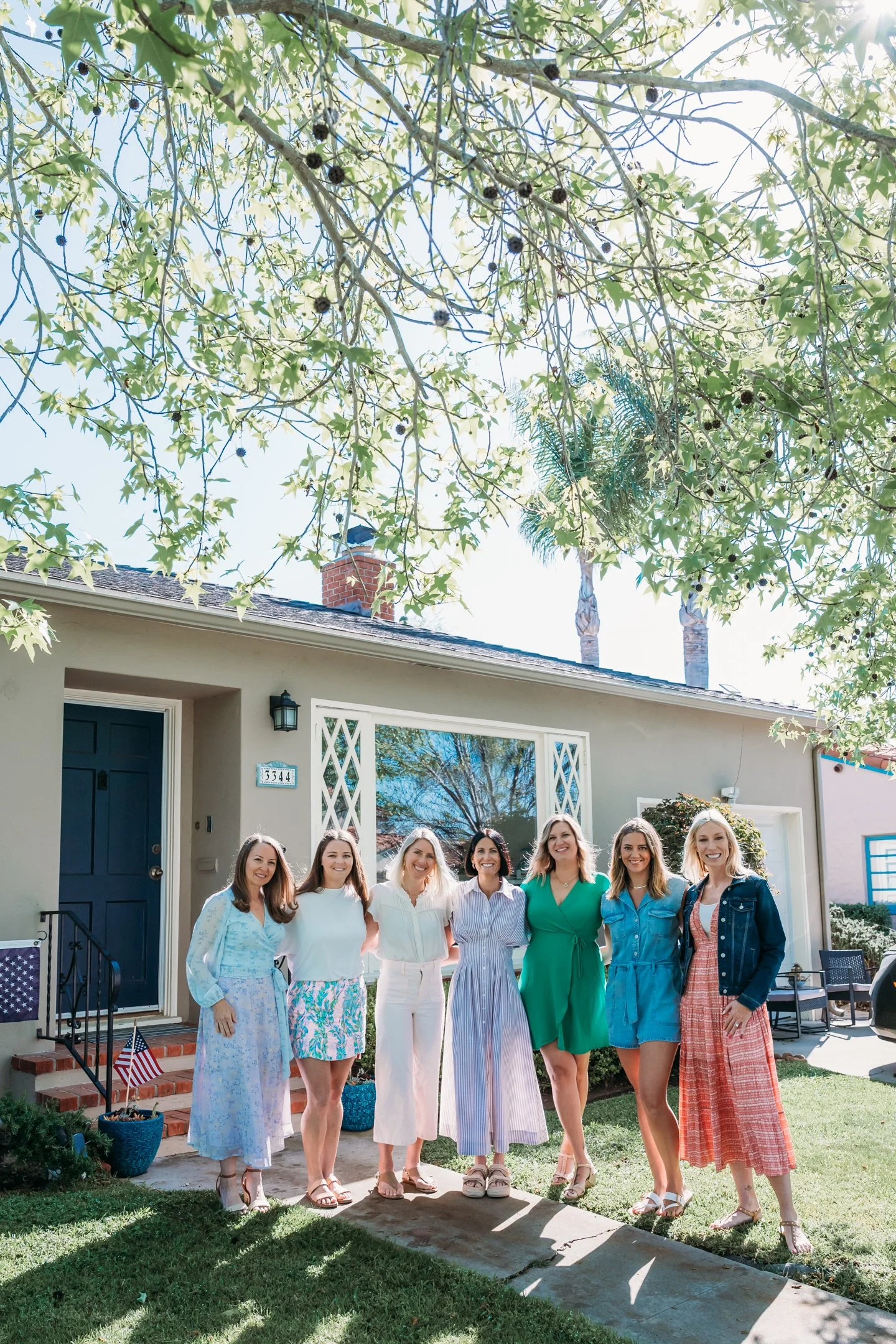 A group of seven women standing on a sidewalk in front of a house, smiling, with a large tree overhead. They are dressed in summer clothing. Alisha Mowry Photography Military Photographer San Diego