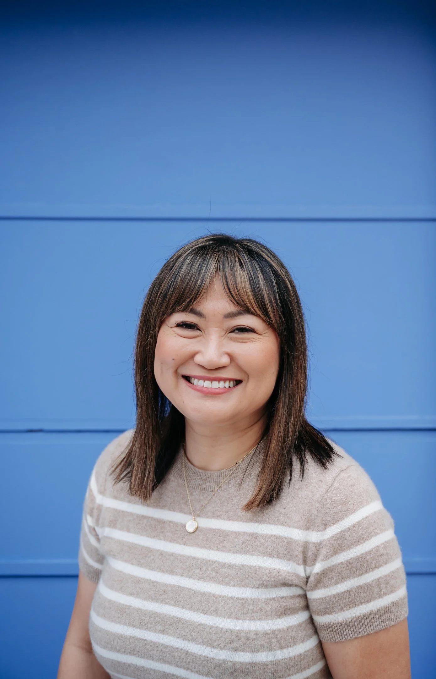 Smiling woman with shoulder-length brown hair in front of a bright blue wall, wearing a beige and white striped shirt and a delicate necklace. Alisha Mowry Photography Military, Brand, and Portrait Photographer San Diego CA