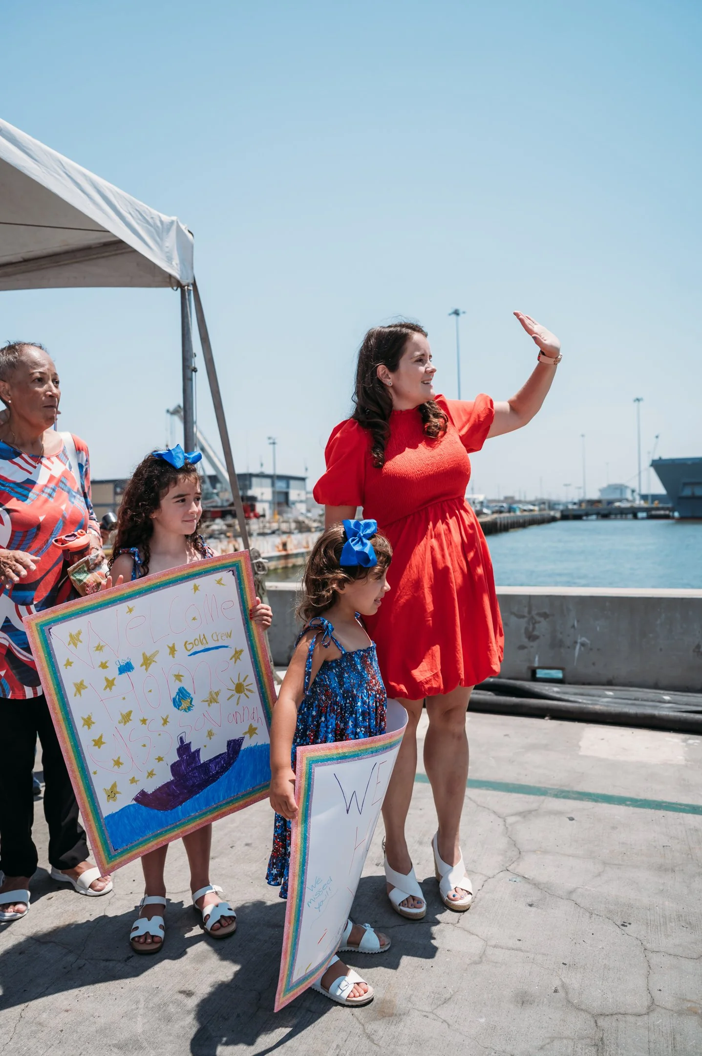 Women and children at a waterfront, one woman in red dress waving, children holding signs with drawings and messages. military homecoming photography in San Diego 
