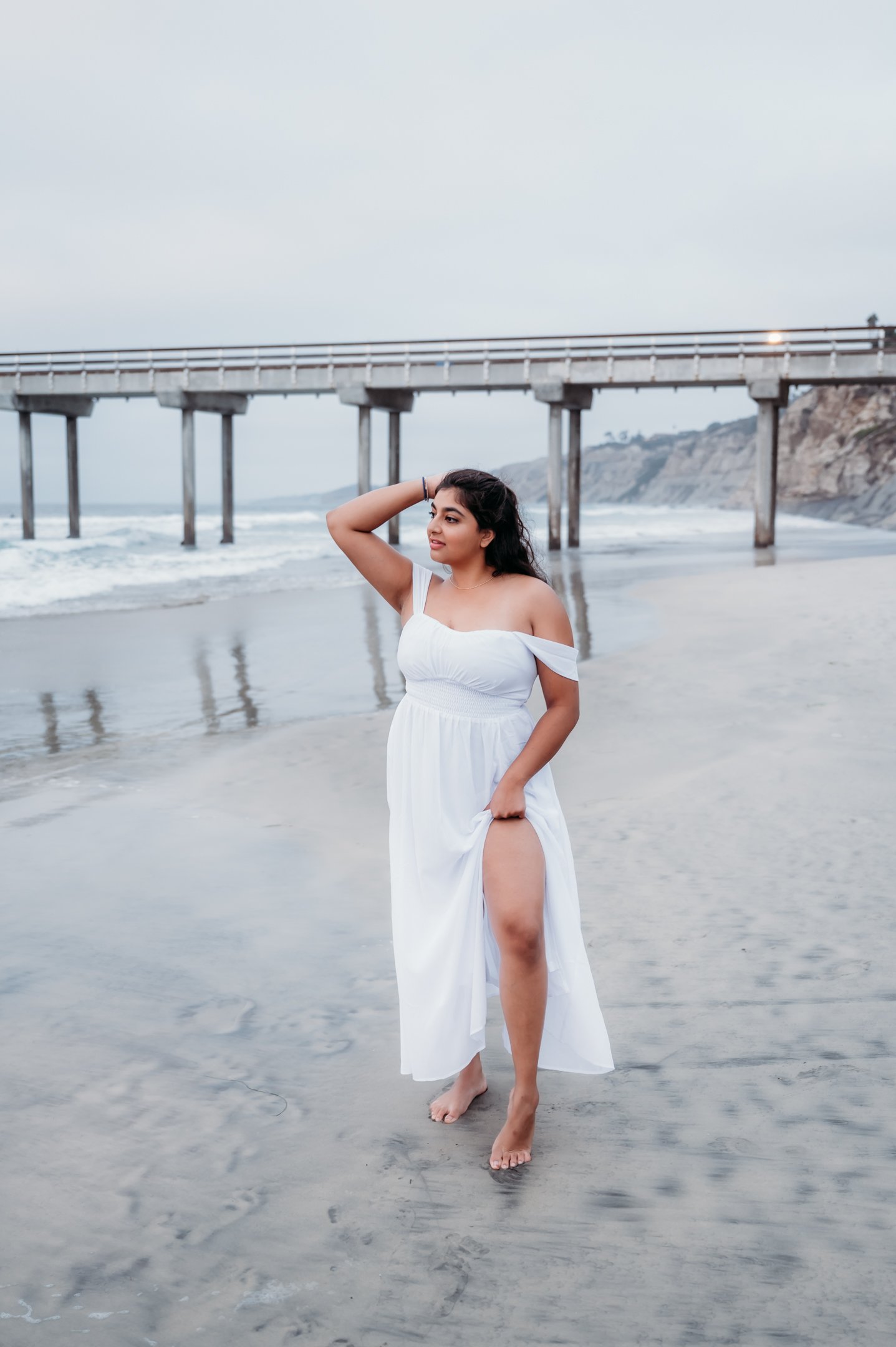 A young woman in a white dress standing barefoot on the beach, holding her dress up to reveal her leg, with a pier in the background under an overcast sky. Alisha Mowry Photography Military, Brand, and Portrait Photographer San Diego CA