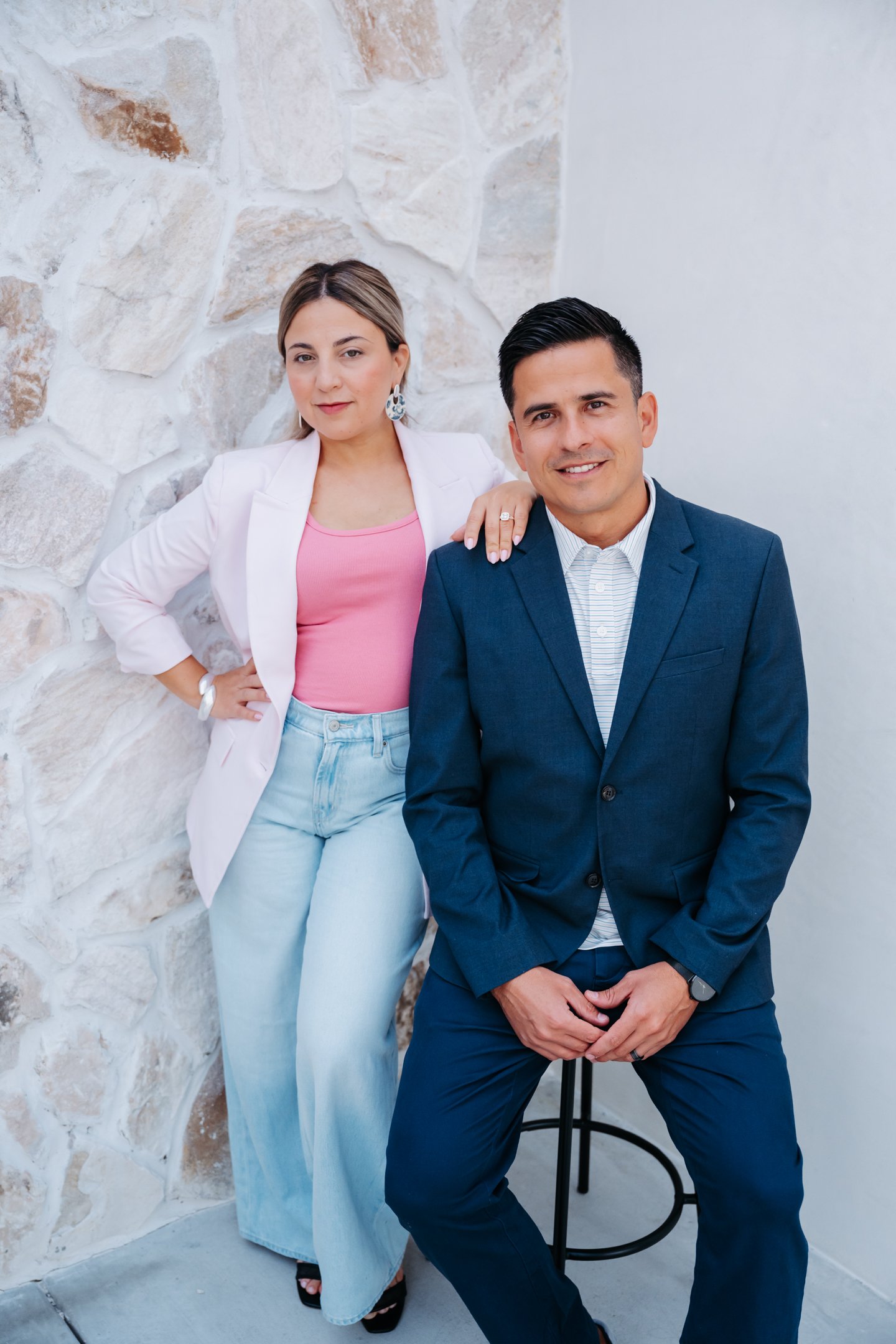 A woman in a white blazer and pink top with light blue jeans standing next to a man in a blue suit, who is sitting on a stool. Alisha Mowry Photography Military, Brand, and Portrait Photographer San Diego CA