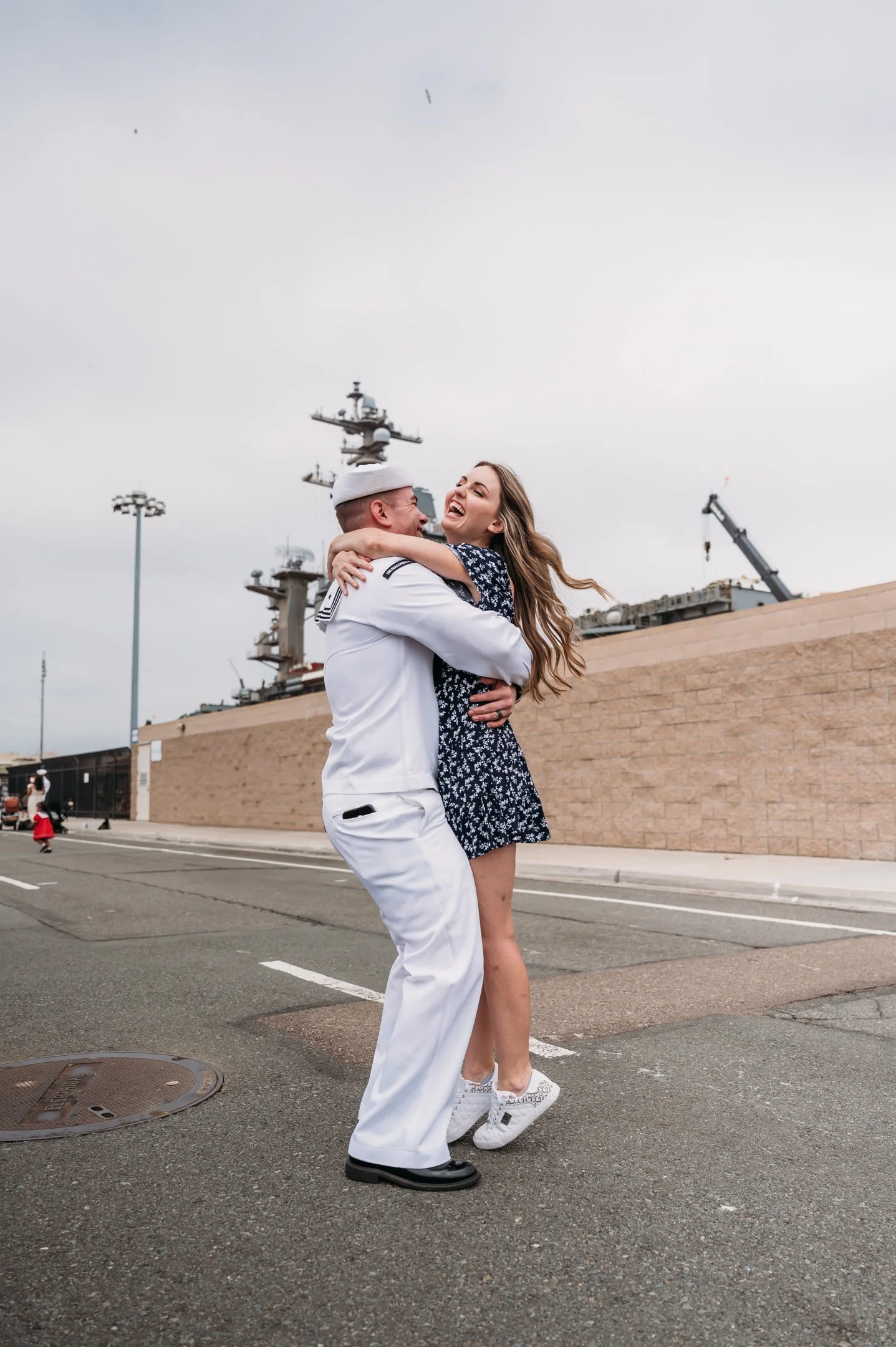 A sailor in uniform dancing with a woman in a blue floral dress on a street near a naval ship, both smiling and embracing each other.   San Diego Military Homecoming Photograph