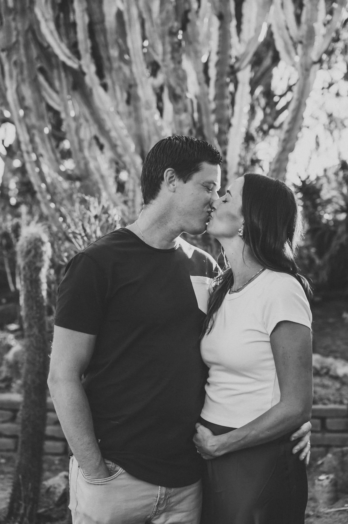 A black and white photo of a couple kissing outdoors, standing close together with a large cactus in the background. Alisha Mowry Photography Military, Brand, and Portrait Photographer San Diego CA
