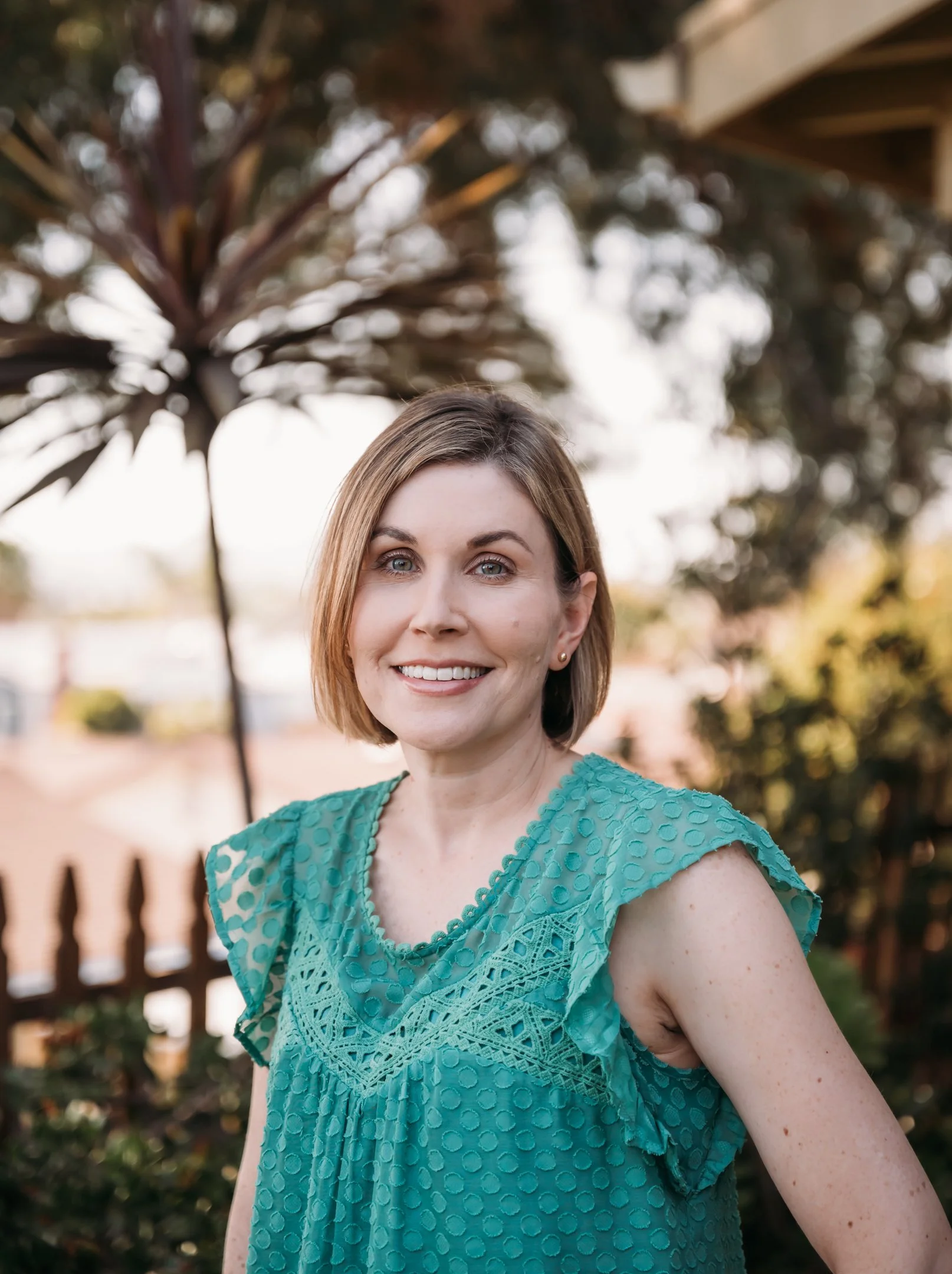 A woman with short brown hair and blue eyes is smiling while standing outdoors in front of trees and a wooden fence, wearing a teal, textured sleeveless top. Alisha Mowry Photography Military, Brand, and Portrait Photographer San Diego CA