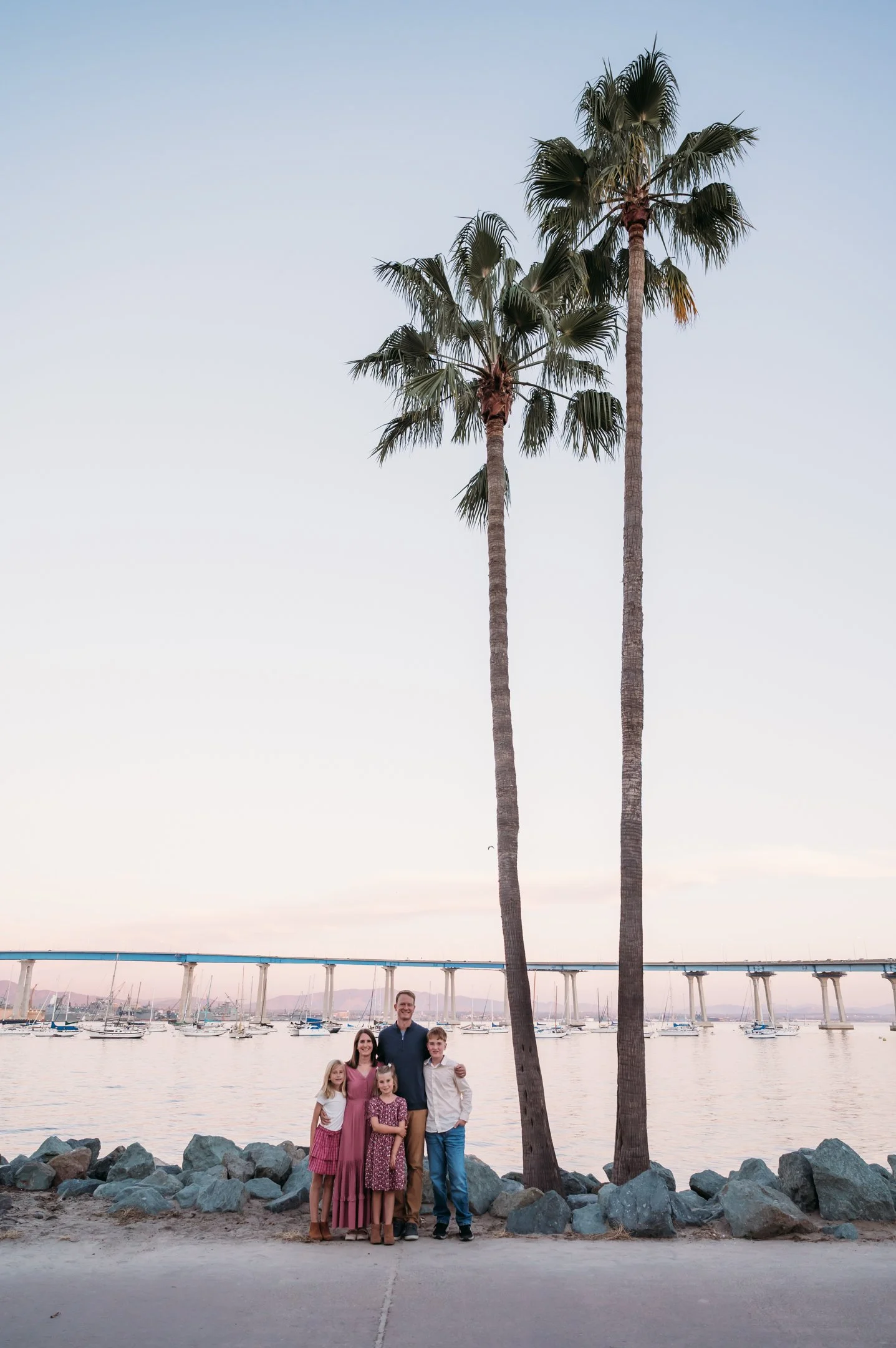 Family of four standing on a lakeside promenade with two palm trees in the background, boats on the water, and a bridge in the distance during sunset. Alisha Mowry Photography Military, Brand, and Portrait Photographer San Diego CA