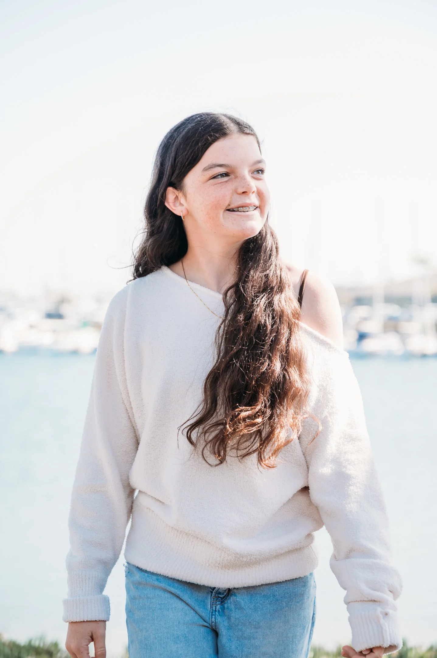 A young girl with long, wavy brown hair standing outdoors near a marina with boats in the background, smiling and looking to the side. Alisha Mowry Photography Military, Brand, and Portrait Photographer San Diego CA