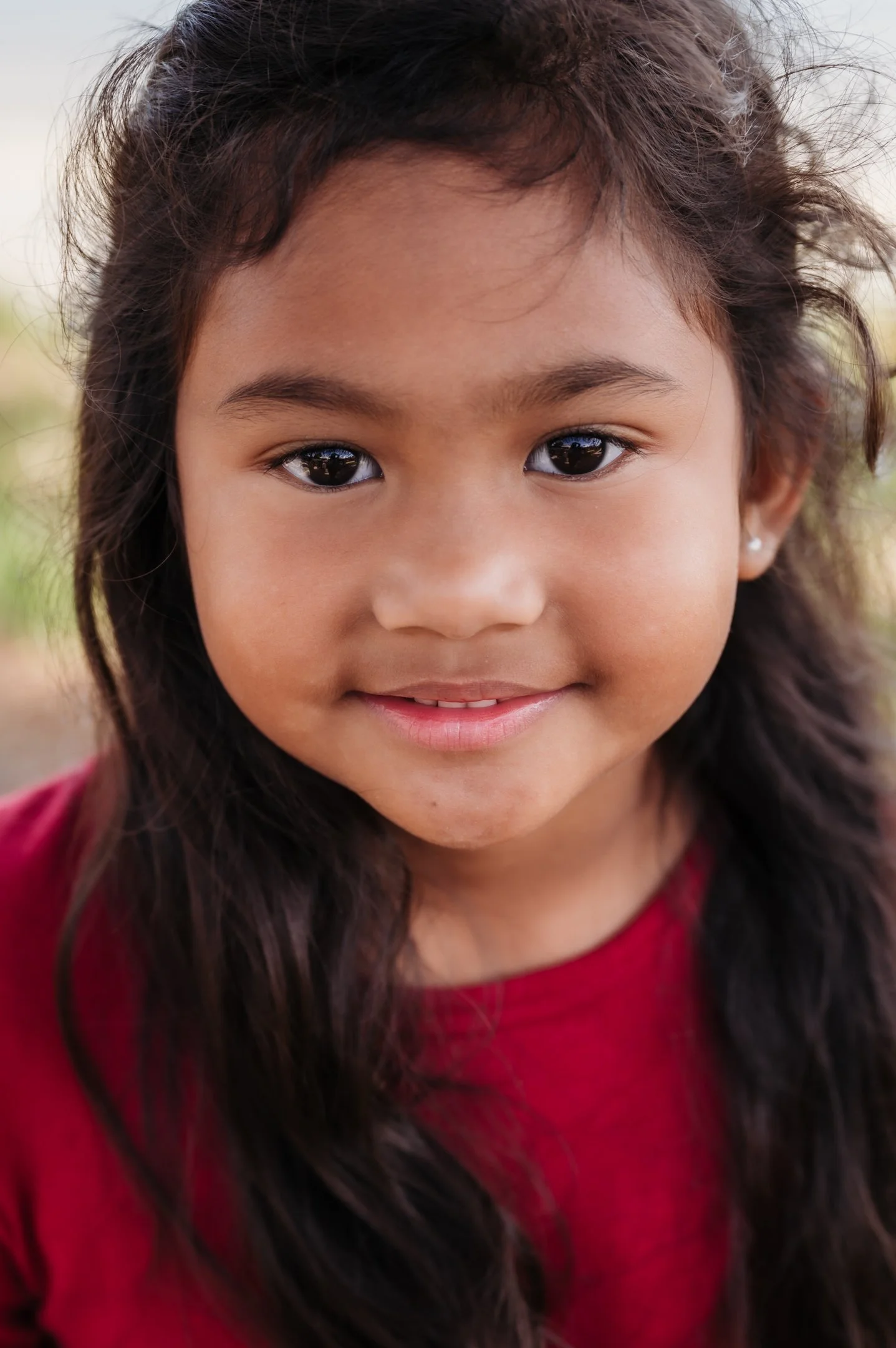 Close-up of a young girl with dark hair, brown eyes, and a slight smile, wearing a red shirt, outdoors with blurred background. Alisha Mowry Photography Military, Brand, and Portrait Photographer San Diego CA