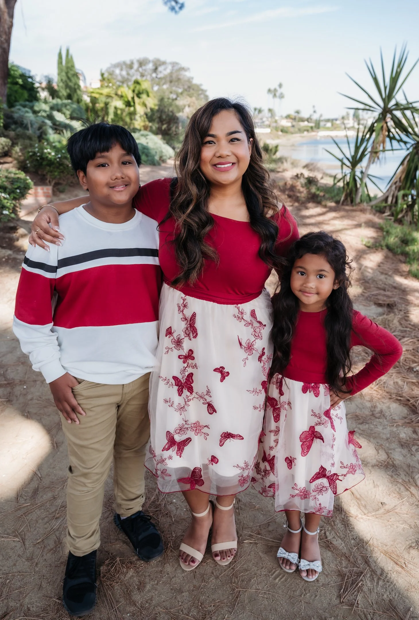 A woman and two children standing outdoors by a body of water, smiling at the camera. They are dressed in coordinated red and white outfits, with butterflies on the girl's dress and the woman's skirt. The background includes trees, bushes, and a sand