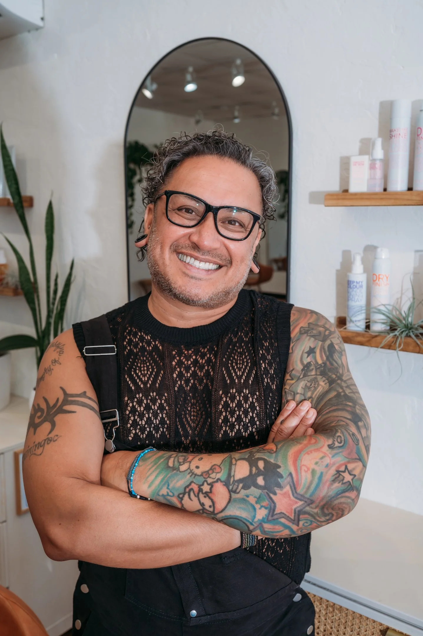 A smiling man with gray curly hair, wearing glasses, a black sleeveless mesh top, and colorful tattoos on his arms, standing inside a room with a mirror and shelves with skincare products. Alisha Mowry Photography Military, Brand, and Portrait Photog